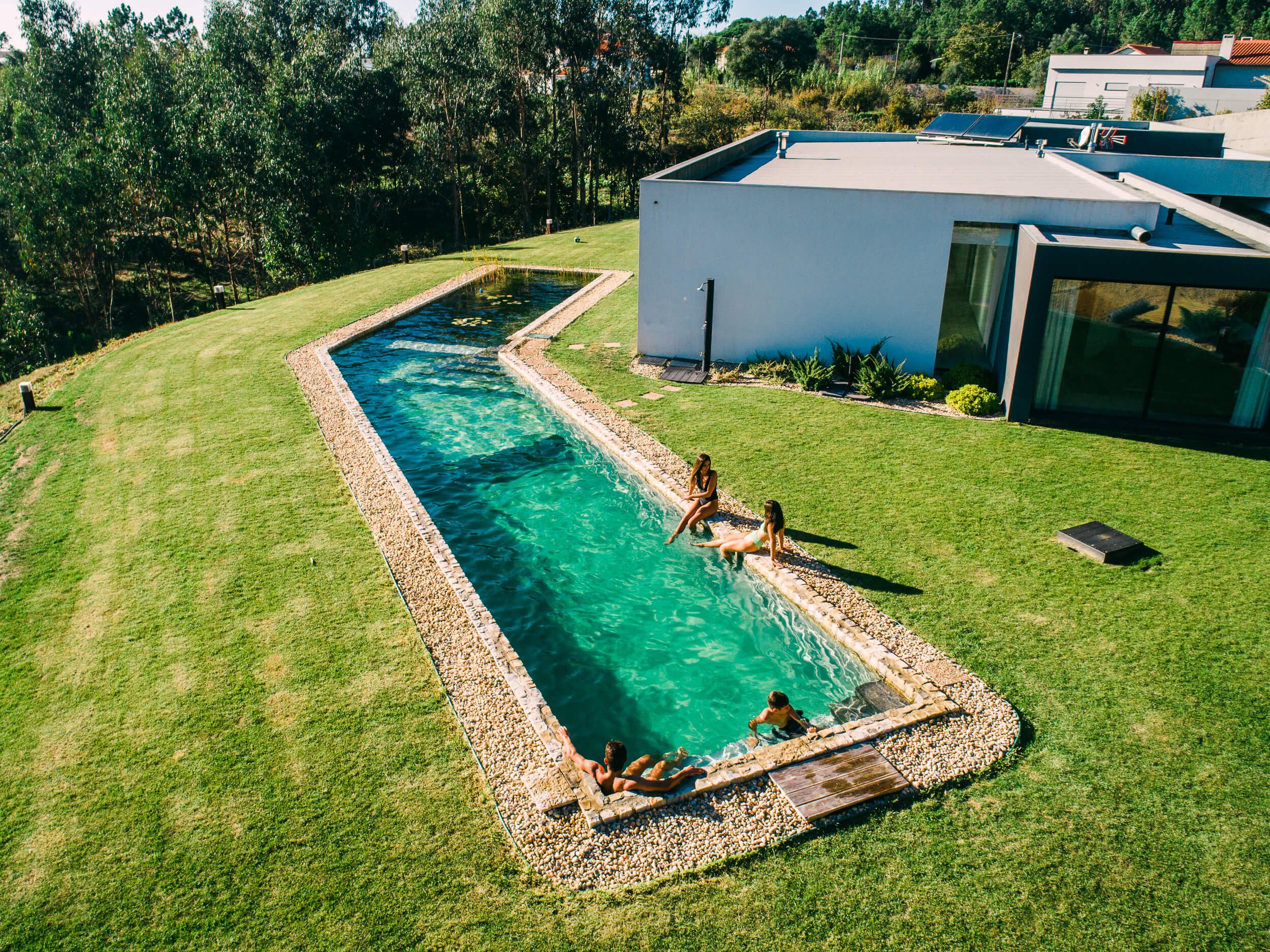 Piscina retangular em um jardim gramado com pessoas relaxando à beira e dentro da água, ao lado de uma casa moderna.