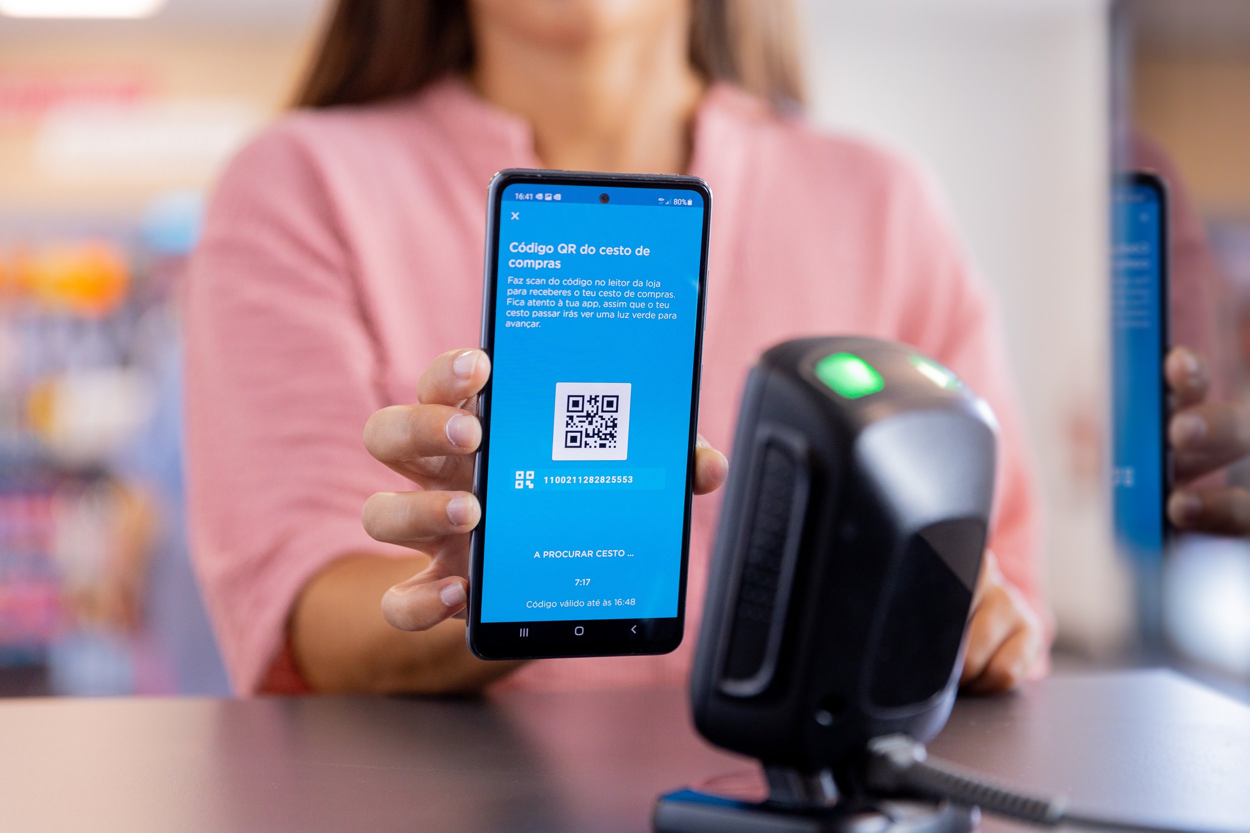 Pessoa segurando um smartphone com um código QR em frente a um leitor de código de barras em uma loja.