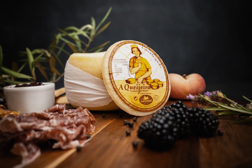 Queijo embalado ao lado de presunto, amoras e ramos de ervas sobre uma tábua de madeira.