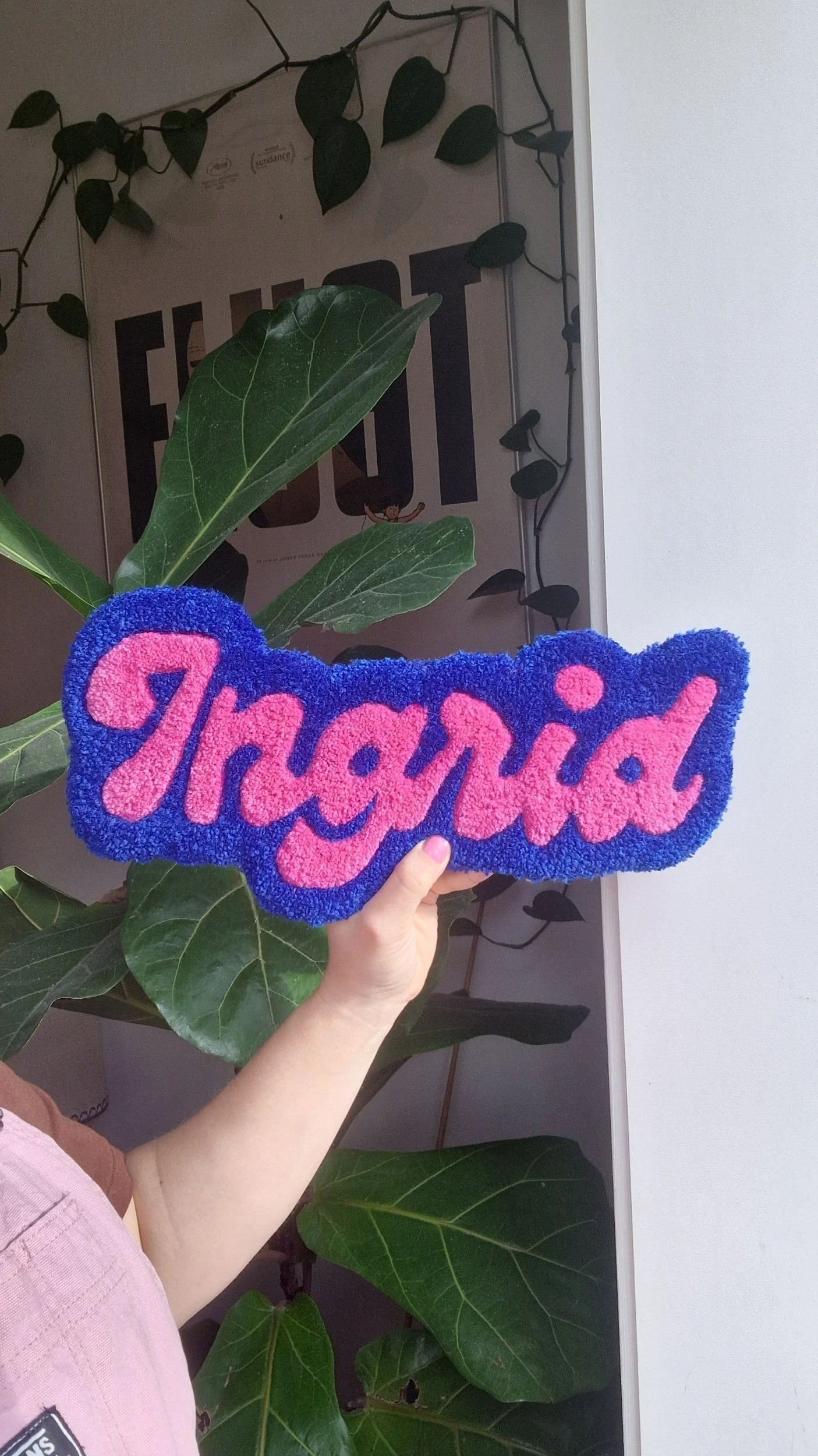 Der holded et personligt tuftet navnetæppe med teksten "Ingrid" i pink og blå. Baggrund med grønne store blade.