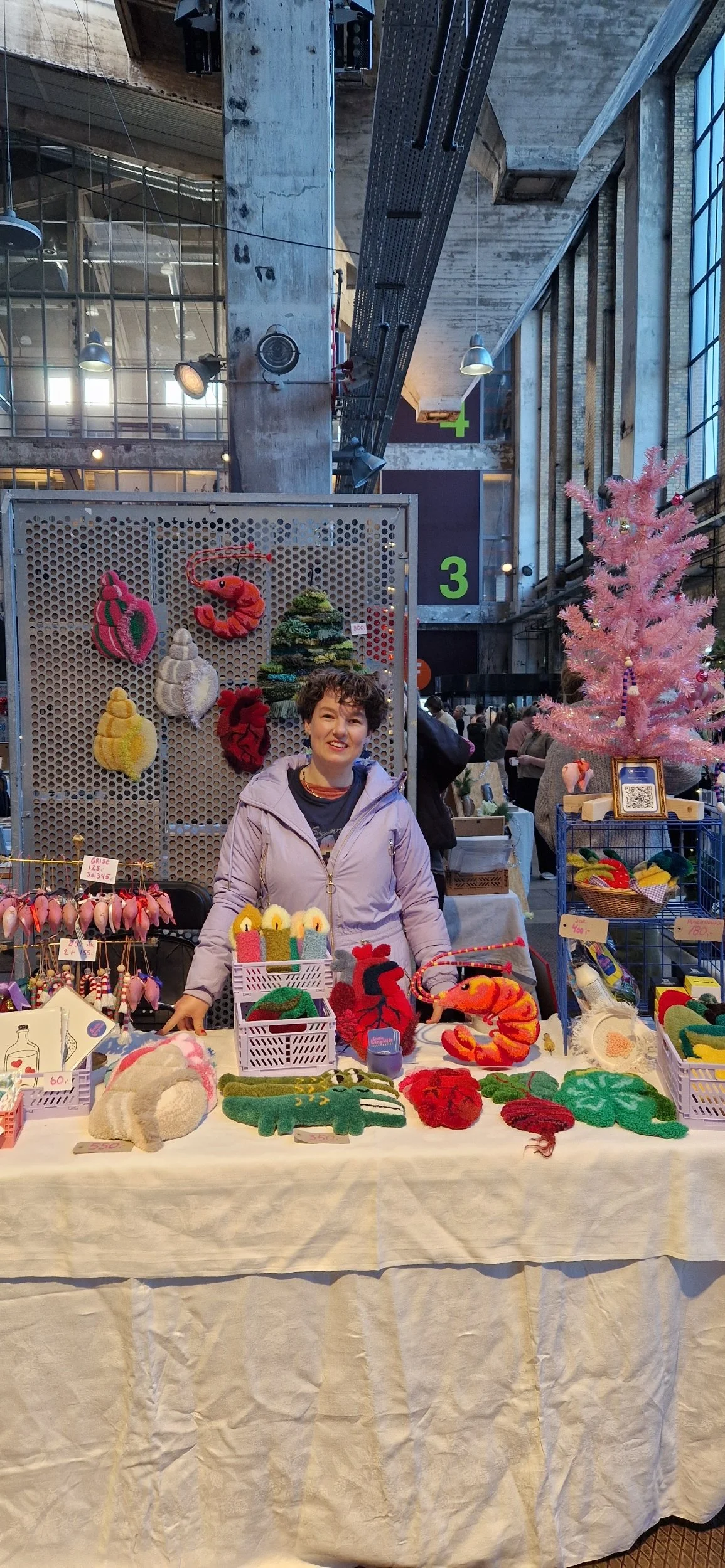 Kunsthåndværker står bag en bod med julepynt, herunder strikkede ornamenter og nissehuer, i en indendørs markedshal med store vinduer og højt til loftet.