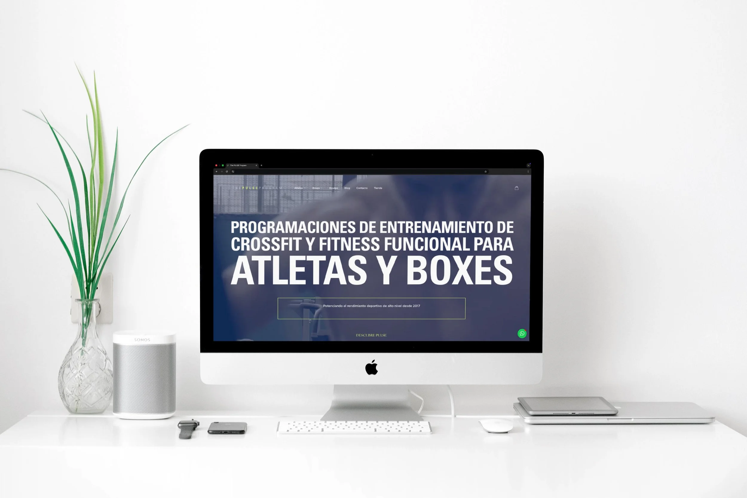 A modern white desk with an iMac displaying a website about CrossFit training programs, a white keyboard and mouse, a tablet, a smartphone, a Sonos speaker, a small device, a glass vase with green plant stems, and a small green WhatsApp icon at the bottom right corner of the screen.