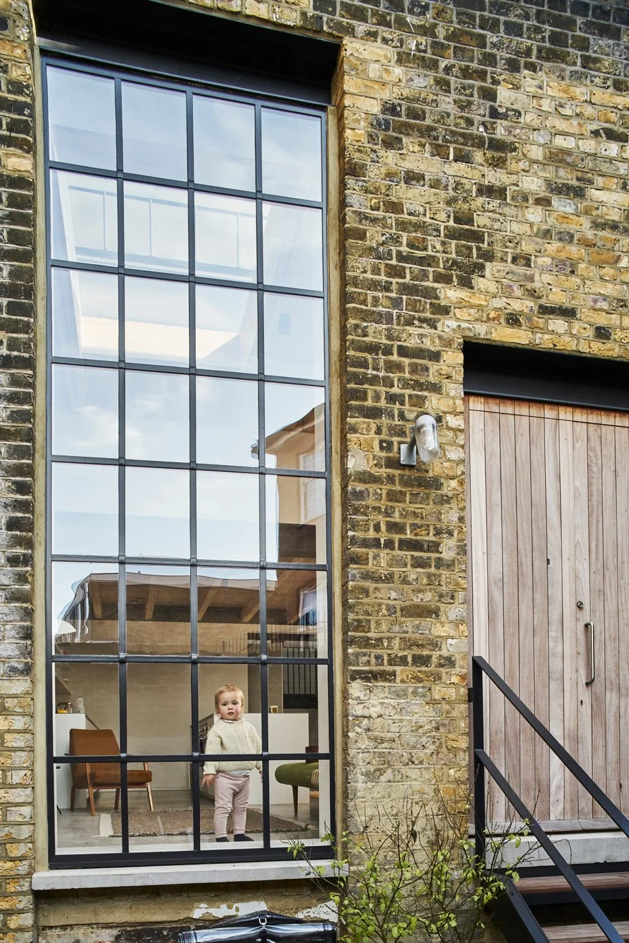 Child looking out of a large critter warehouse window in a contemporary converted Victorian Gin Distillery family house in East London. The front door is iroko timber and the facade is London stock brick architecture