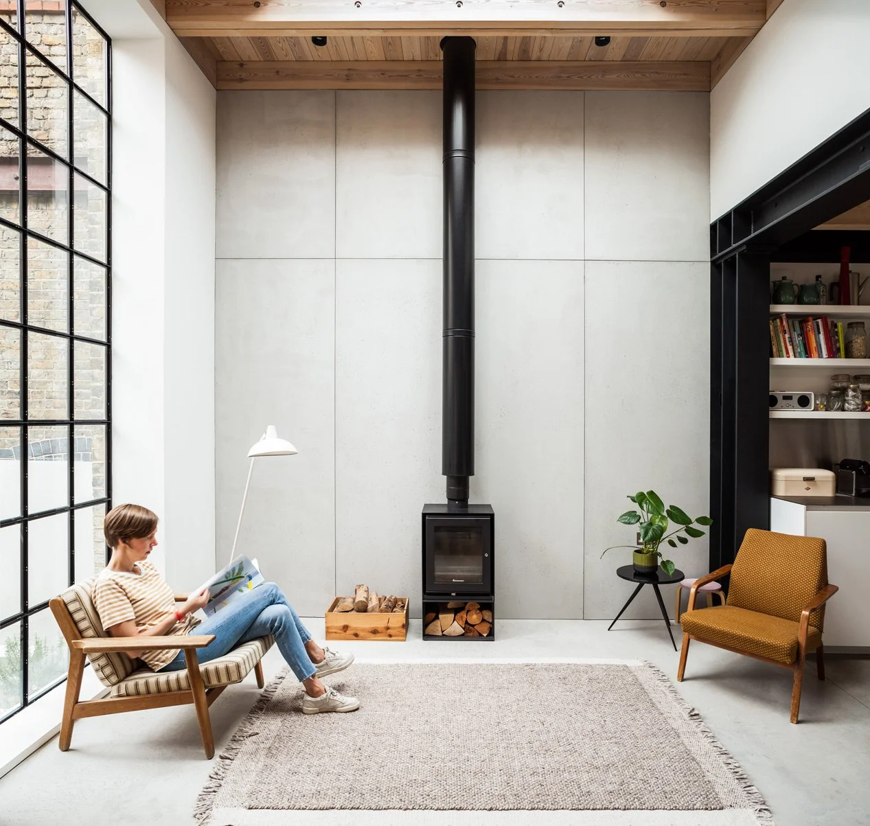 A woman sitting and reading in a contemporary living room with high ceilings, light grey concrete-panelled walls, polished concrete grey floor, full-height black crittall window and a central black wood burner and flue with mid-century furniture