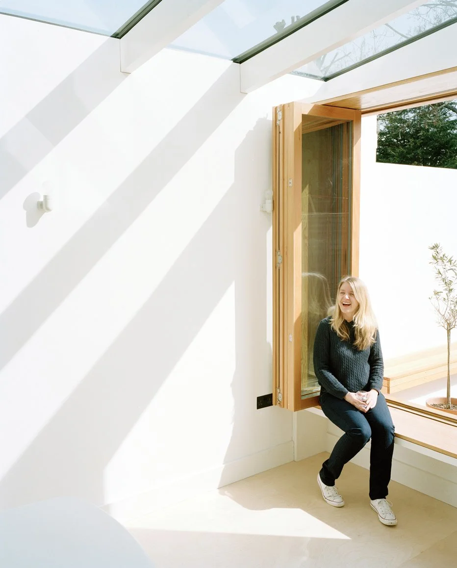 Light-filled white-painted room with a light floor and a glass and timber joist roof with a timber tri-fold window opening onto a courtyard garden with a blonde smiling woman wearing navy blue sitting on the sill