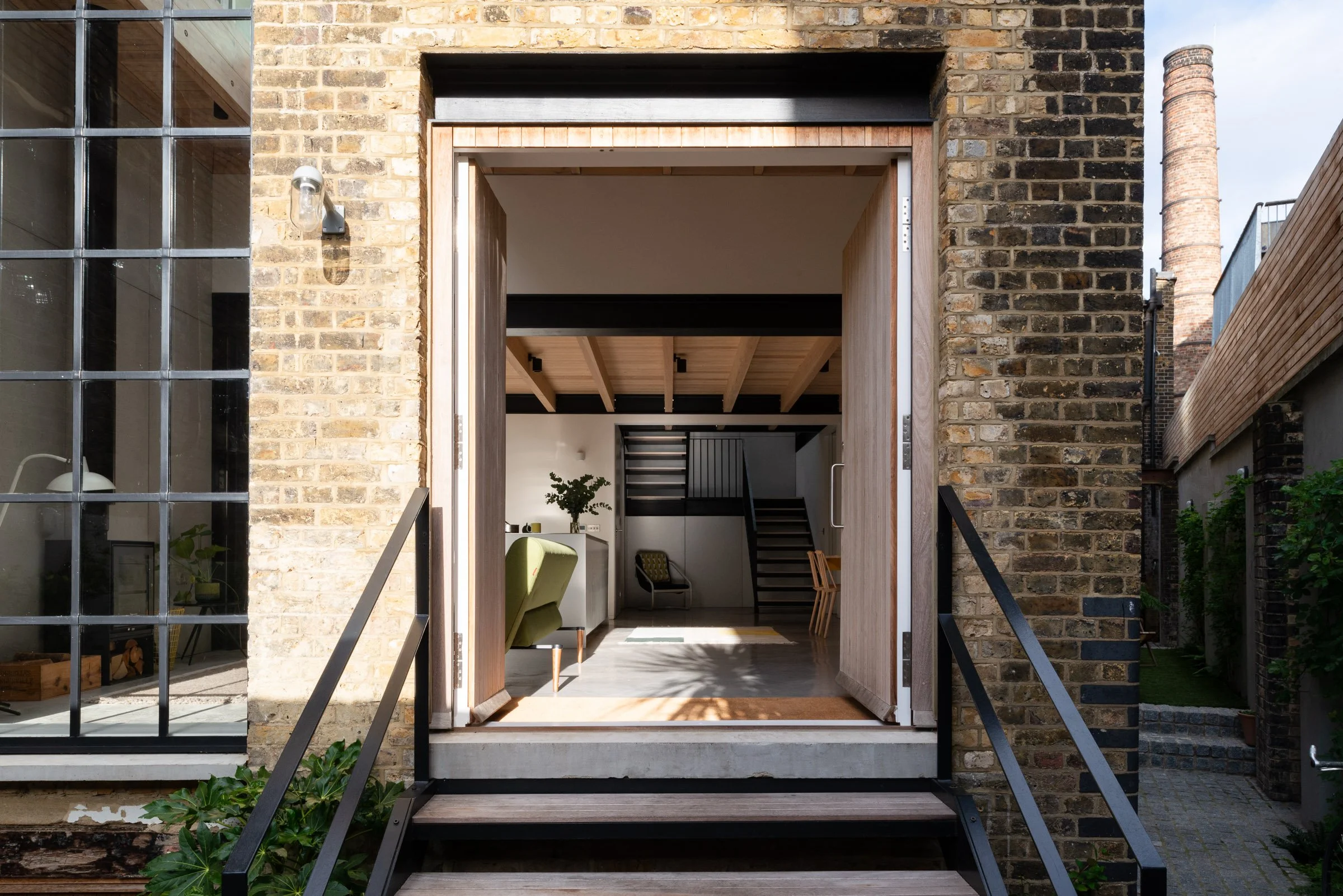 Contemporary renovation of a Victorian Gin Distillery in East London with London stock brick and a white-stained iroko timber door opening onto a contemporary light natural interior with exposed timber joists and black steel features