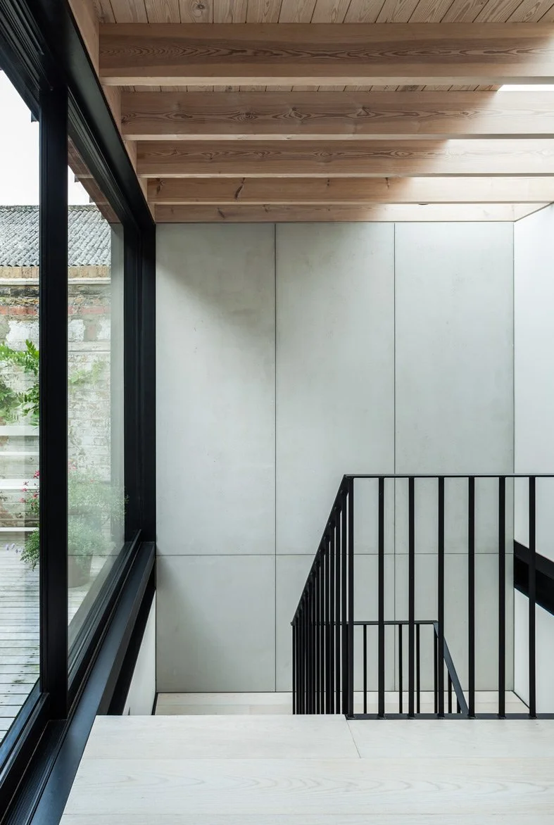 Light flooding in to contemporary Victorian Gin Distillery conversion in East London with precast concrete walls, exposed timber joist ceiling and black steel staircase balustrade with a polished concrete floor