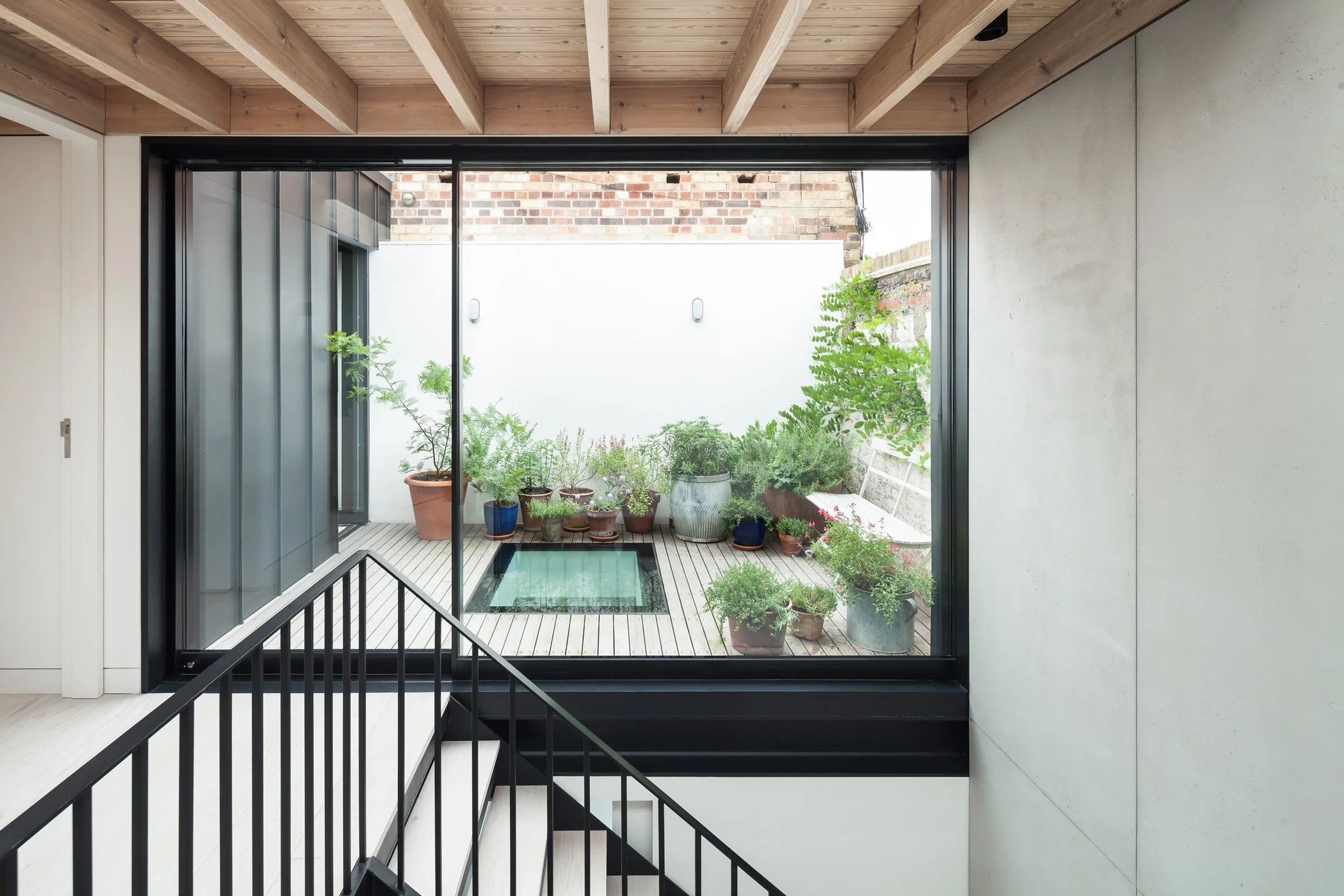 Light flooding in to contemporary Victorian Gin Distillery conversion in East London with precast concrete walls, exposed timber joist ceiling and black steel staircase balustrade with a polished concrete floor. A planted roof terrace draws light in
