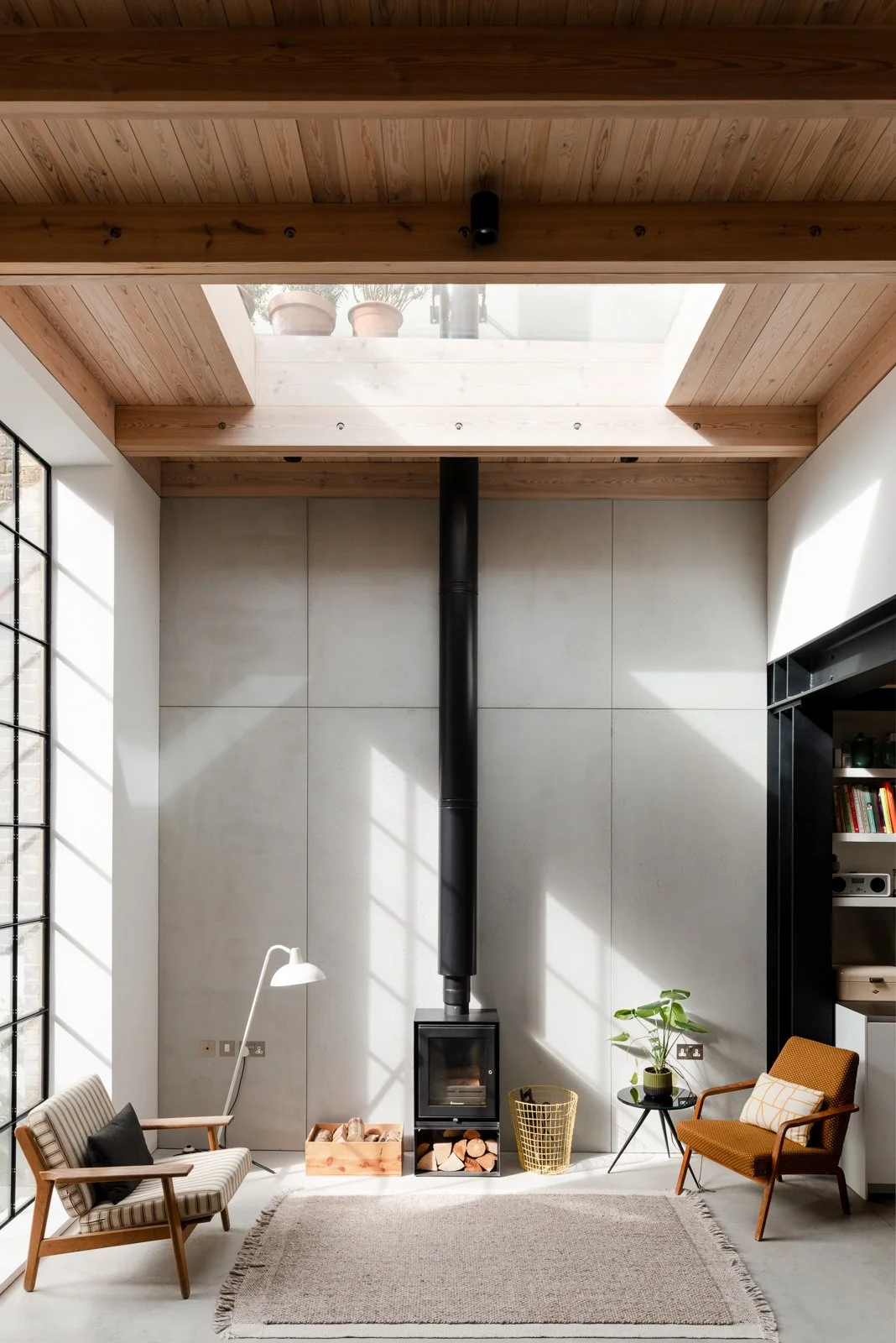 A contemporary living room with high timber ceilings, light grey concrete-panelled walls, polished concrete grey floor, full-height black crittall window and a central black wood burner and flue with mid-century furniture
