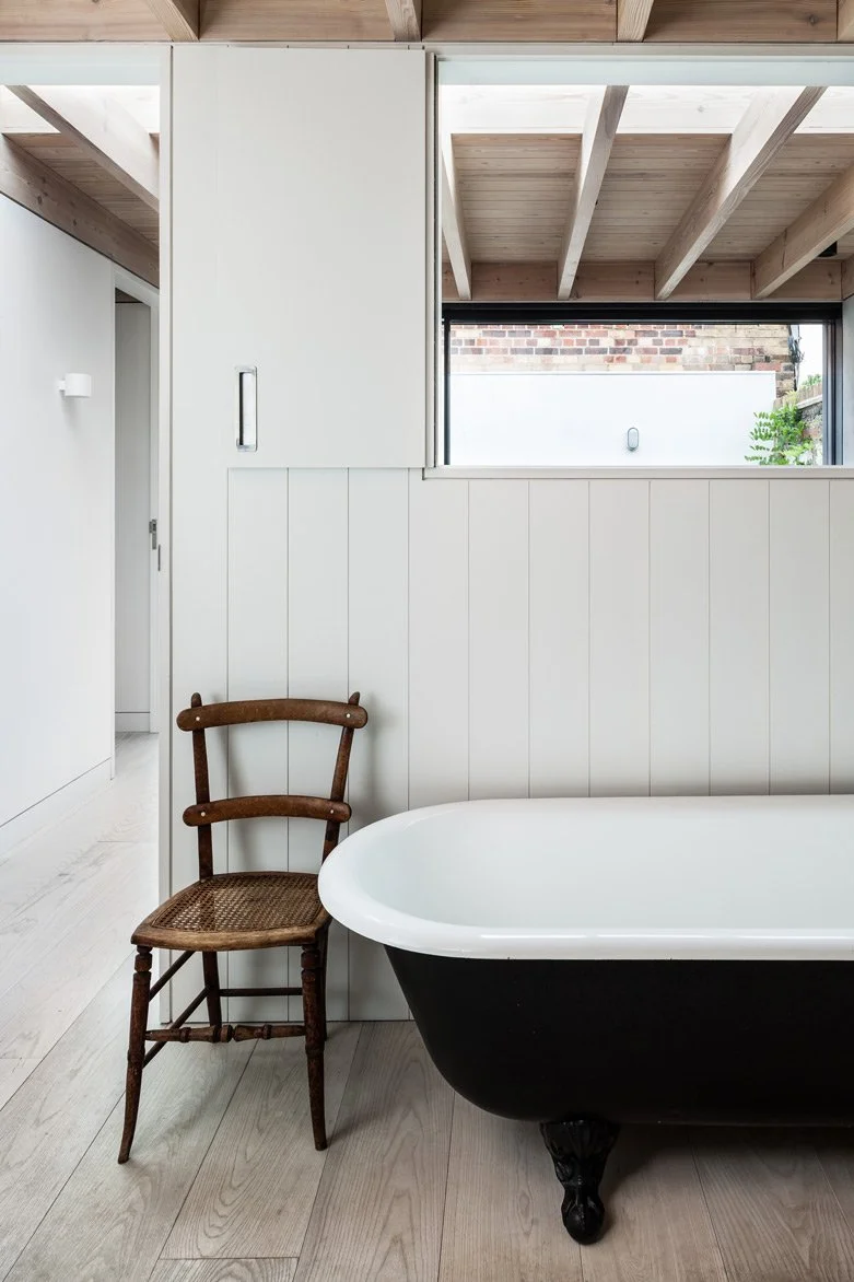 White timber panelled bathroom with a shutter looking through the bedroom with exposed timber ceiling joists to a courtyard with vertical plants growing up the walls. Natural timber furniture and traditional bath visible