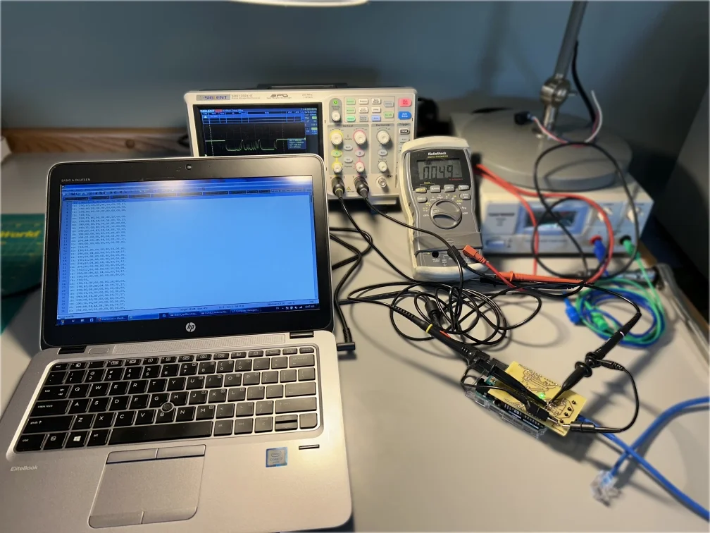 Electronics workbench with a laptop, an oscilloscope, a multimeter, and a circuit board on a gray work surface.