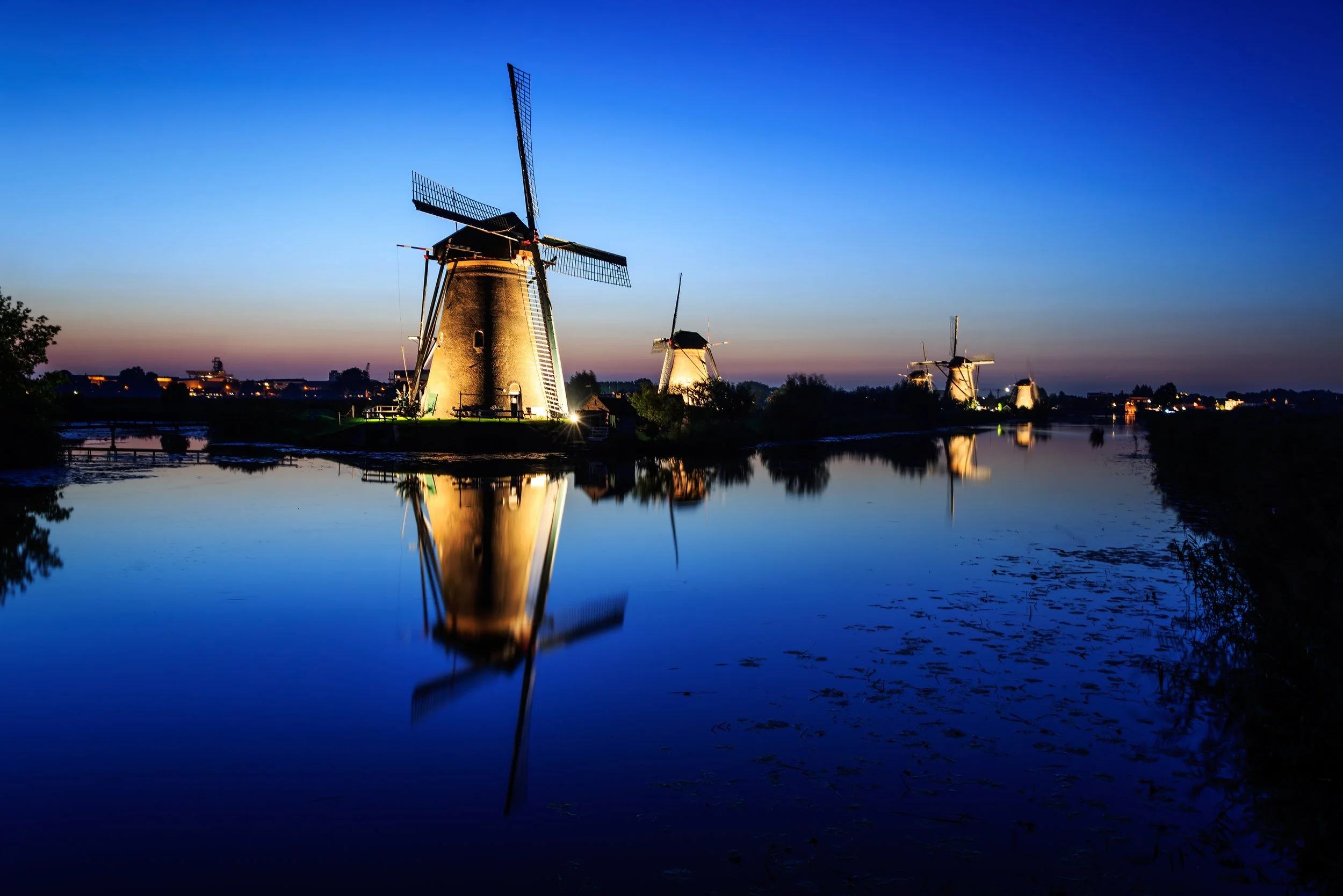 Illuminated windmills reflected in calm water at dusk under a deep blue sky.