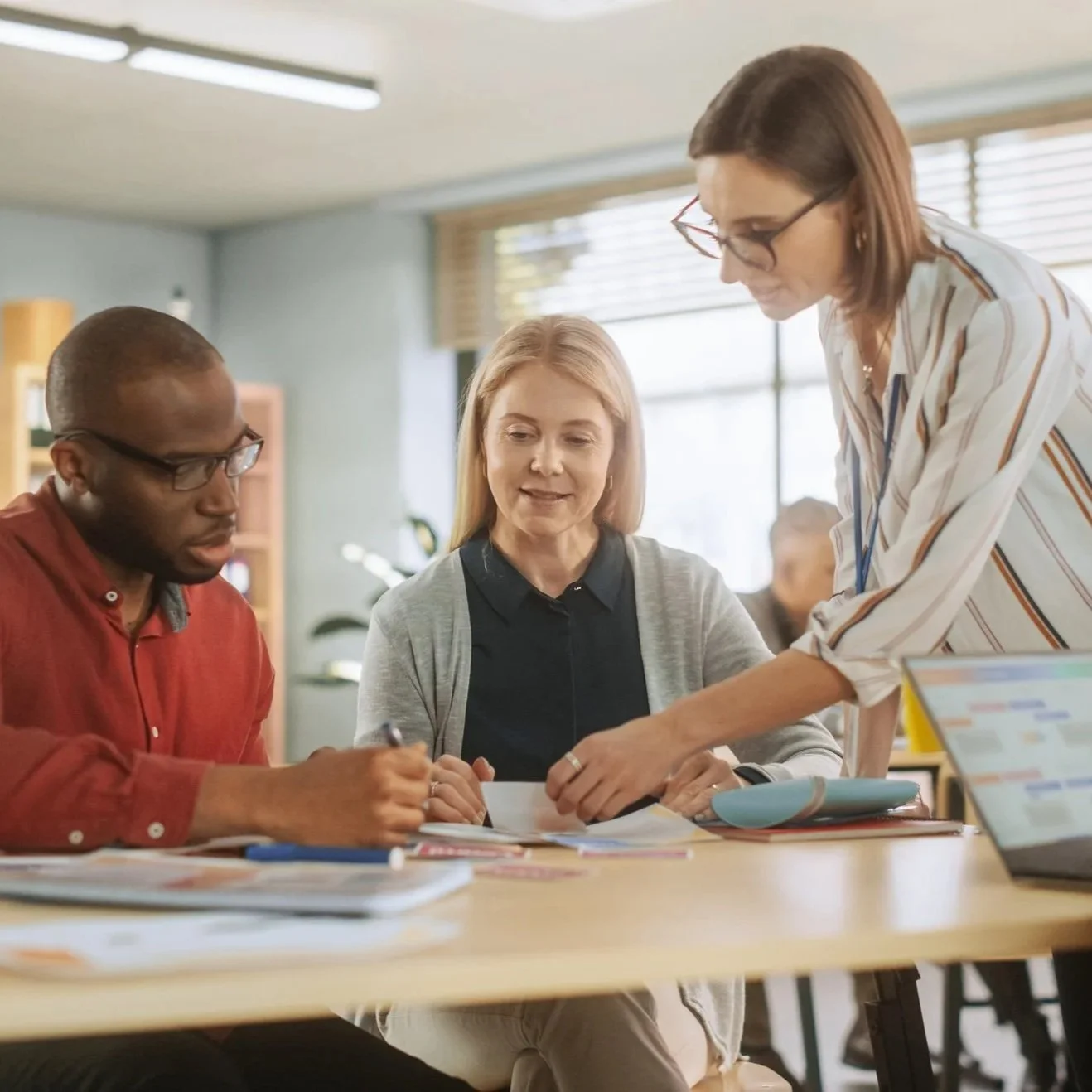 Employees collaborating at a table in a workplace setting, reviewing documents together while one person points to materials and others take notes.