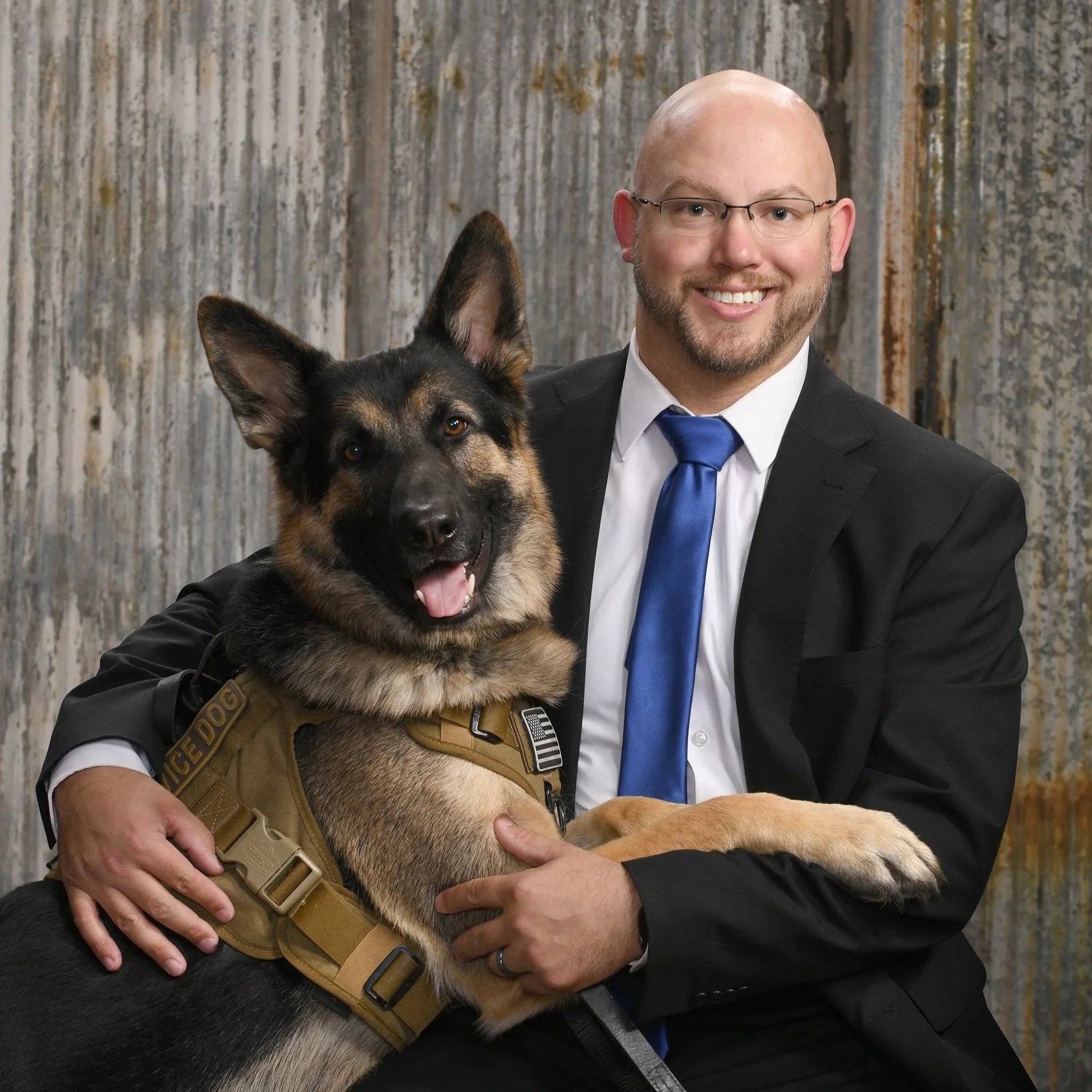 Tony Russell, wearing a black suit and blue tie, sits smiling while holding a German Shepherd service dog wearing a working harness, posed in front of a corrugated metal backdrop.