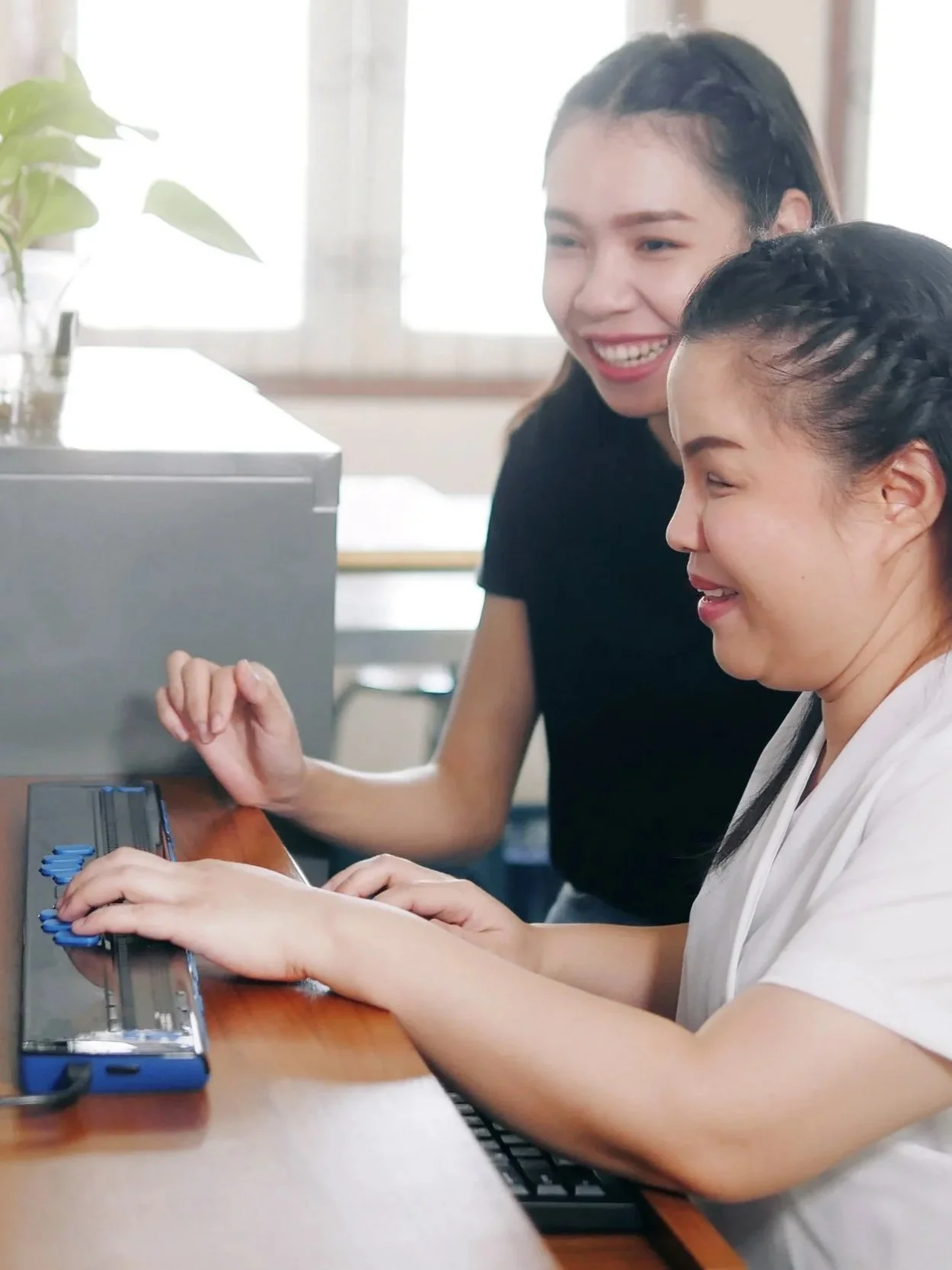 Two coworkers at a computer, one using a braille keyboard with support from a colleague.