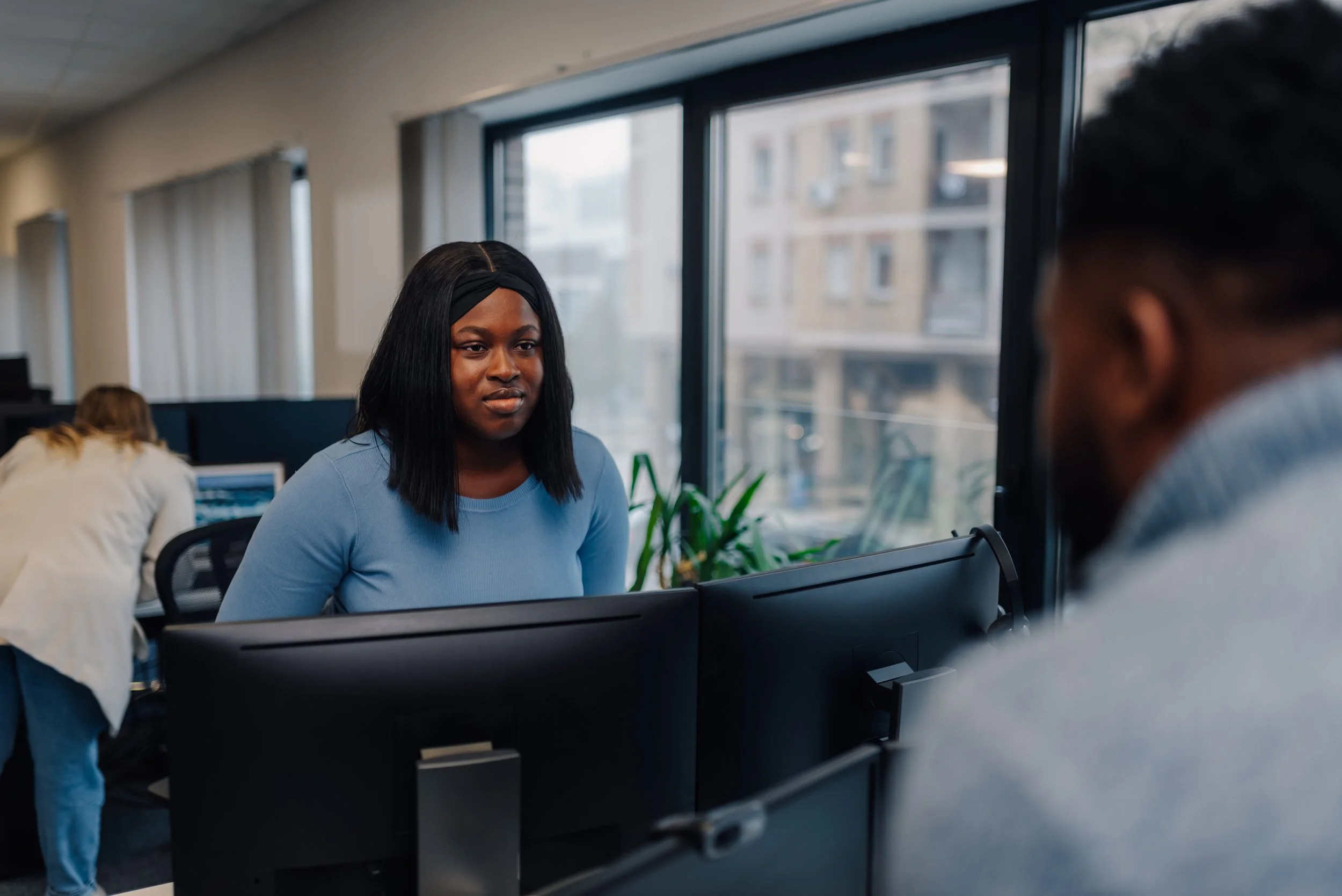Employee working at a dual-monitor workstation in an office, engaged in conversation with a colleague across the desk.