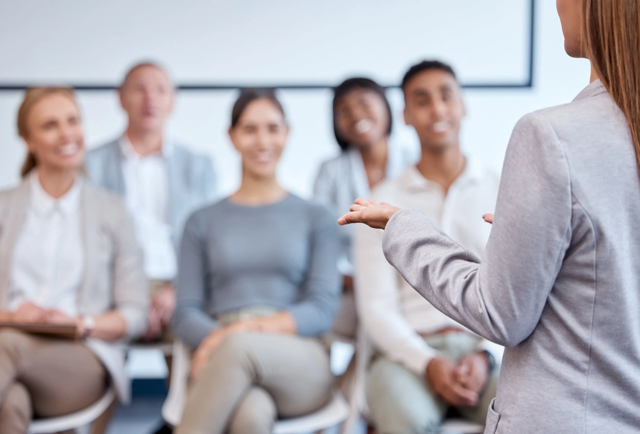 A woman in a gray blazer speaking to a diverse group of five seated professionals, smiling, in a corporate or conference room setting.