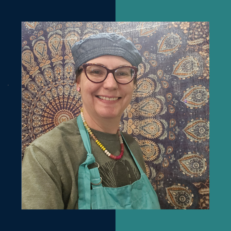 Photo of Shannon Ward, co-founder of Good NAtured, smiling and wearing a gray cap and teal apron in front of a patterned backdrop.