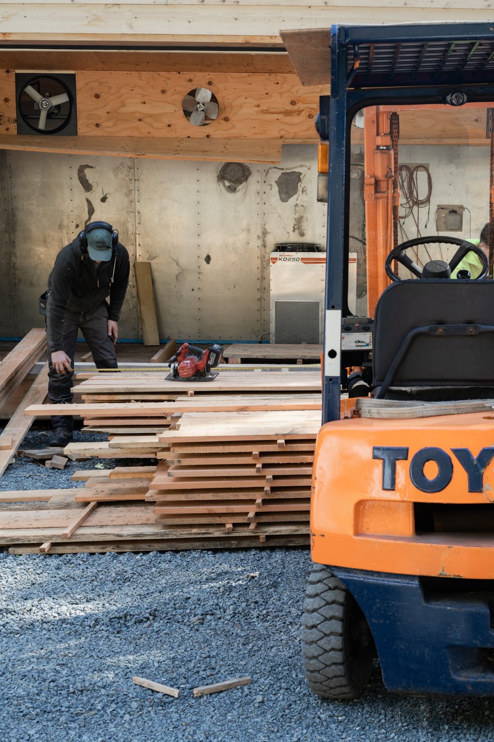 Construction worker in black clothing and a blue cap working on wooden planks inside a building site, with an orange forklift in the foreground and tools like a saw on the wooden pile.
