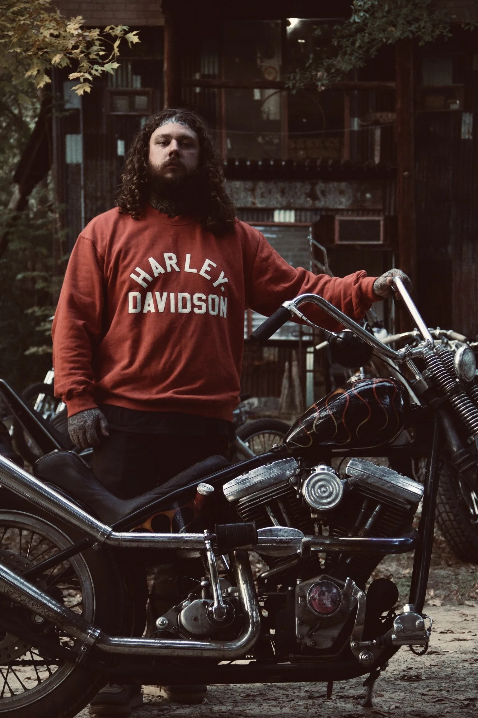 Tattooed man with long curly hair standing beside a vintage custom chopper motorcycle, wearing a red Harley-Davidson sweatshirt in a moody outdoor portrait with a dark film photography aesthetic