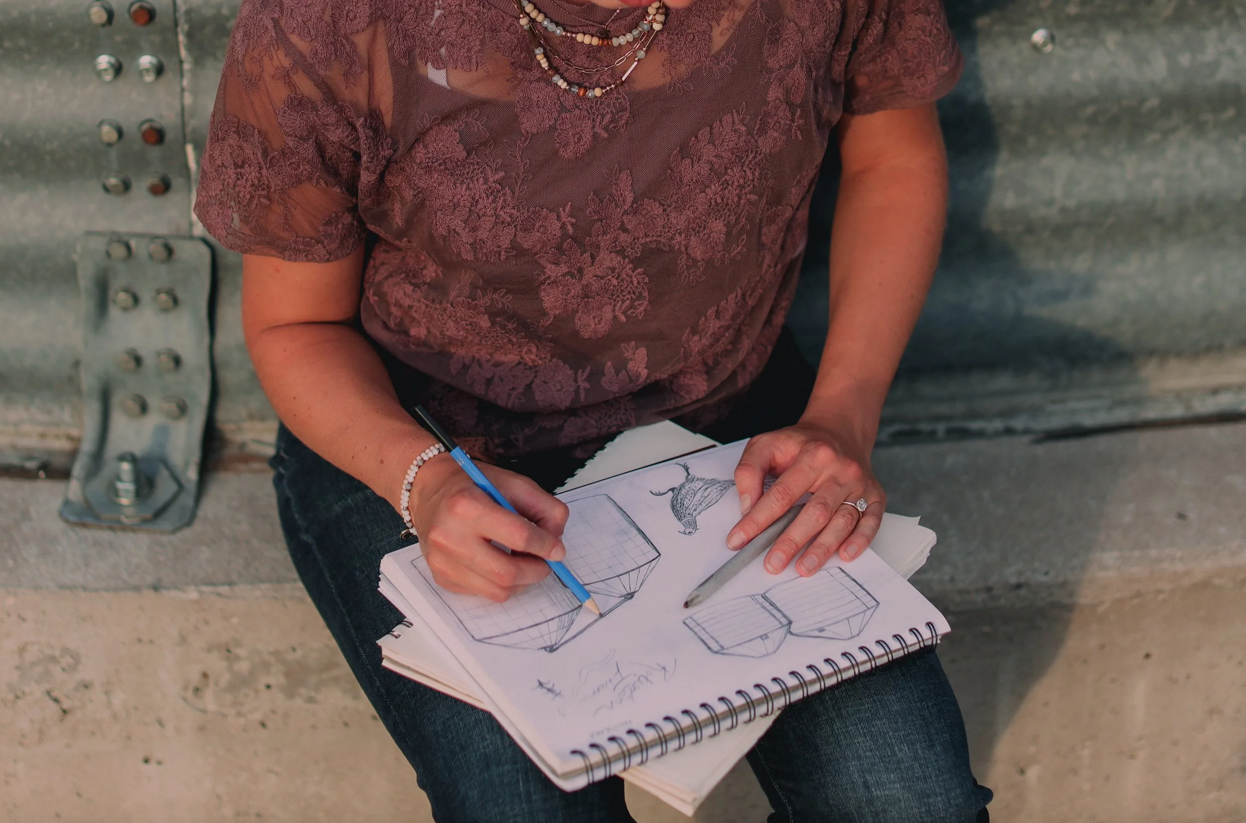 Close-up of a woman sitting on a bench, sketching geometric box designs in a notebook. She is wearing a maroon lace top and layered necklaces, with a silver bracelet on her wrist.
