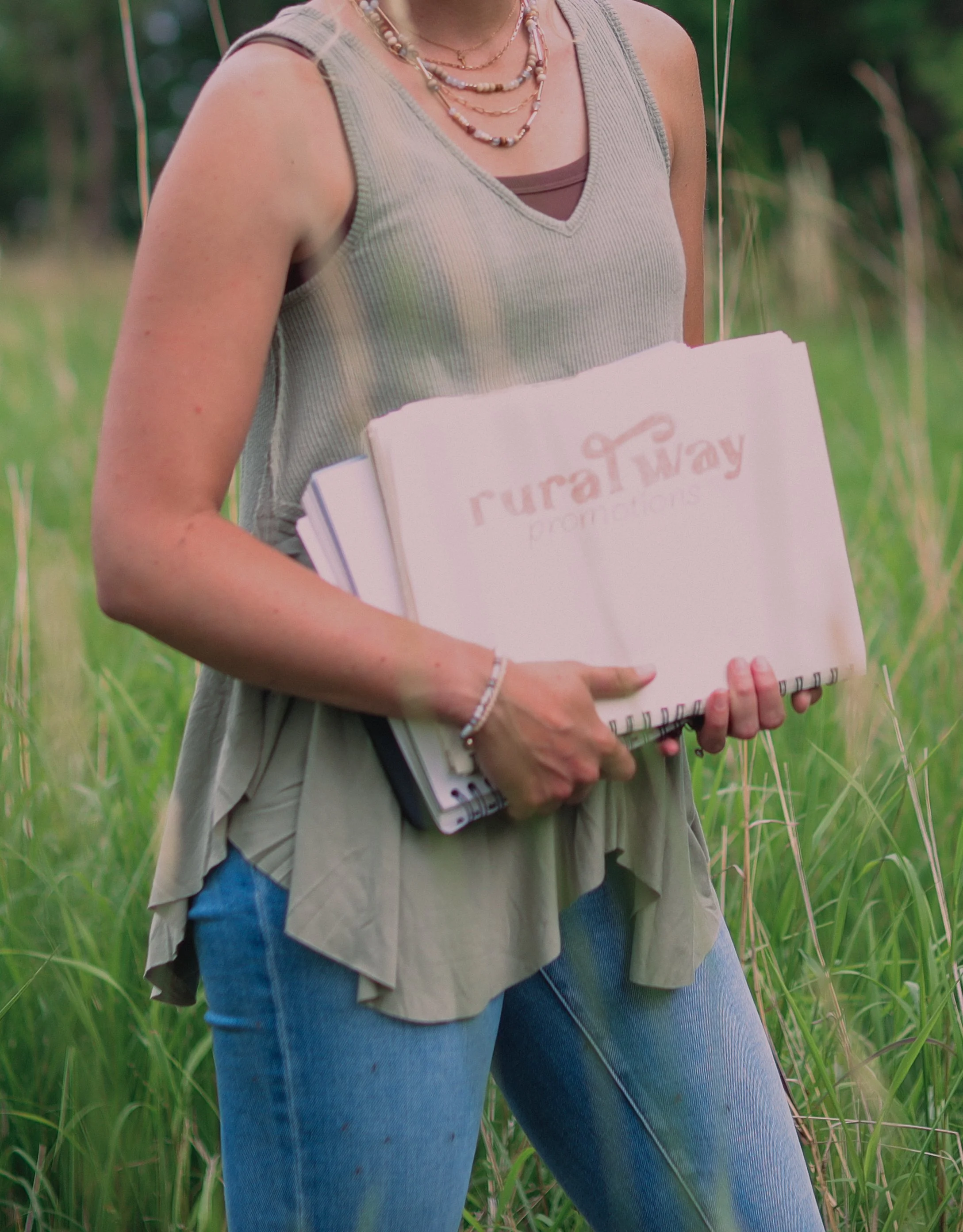 Person holding notebooks standing in tall grass, wearing a sleeveless top, blue jeans, and jewelry, with greenery in the background.