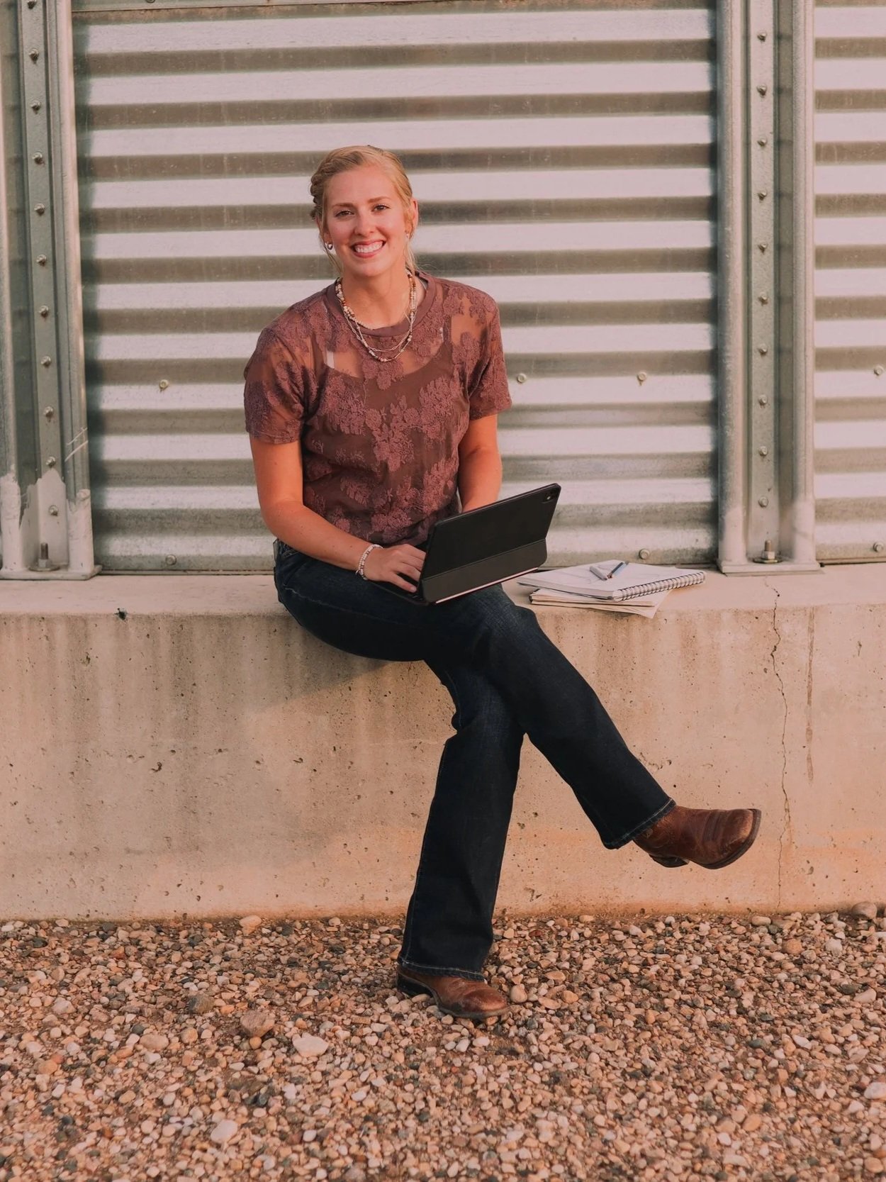 A woman sitting on a concrete ledge with a laptop on her lap, smiling, with notebooks and a pen beside her, outside near a grated metal wall.