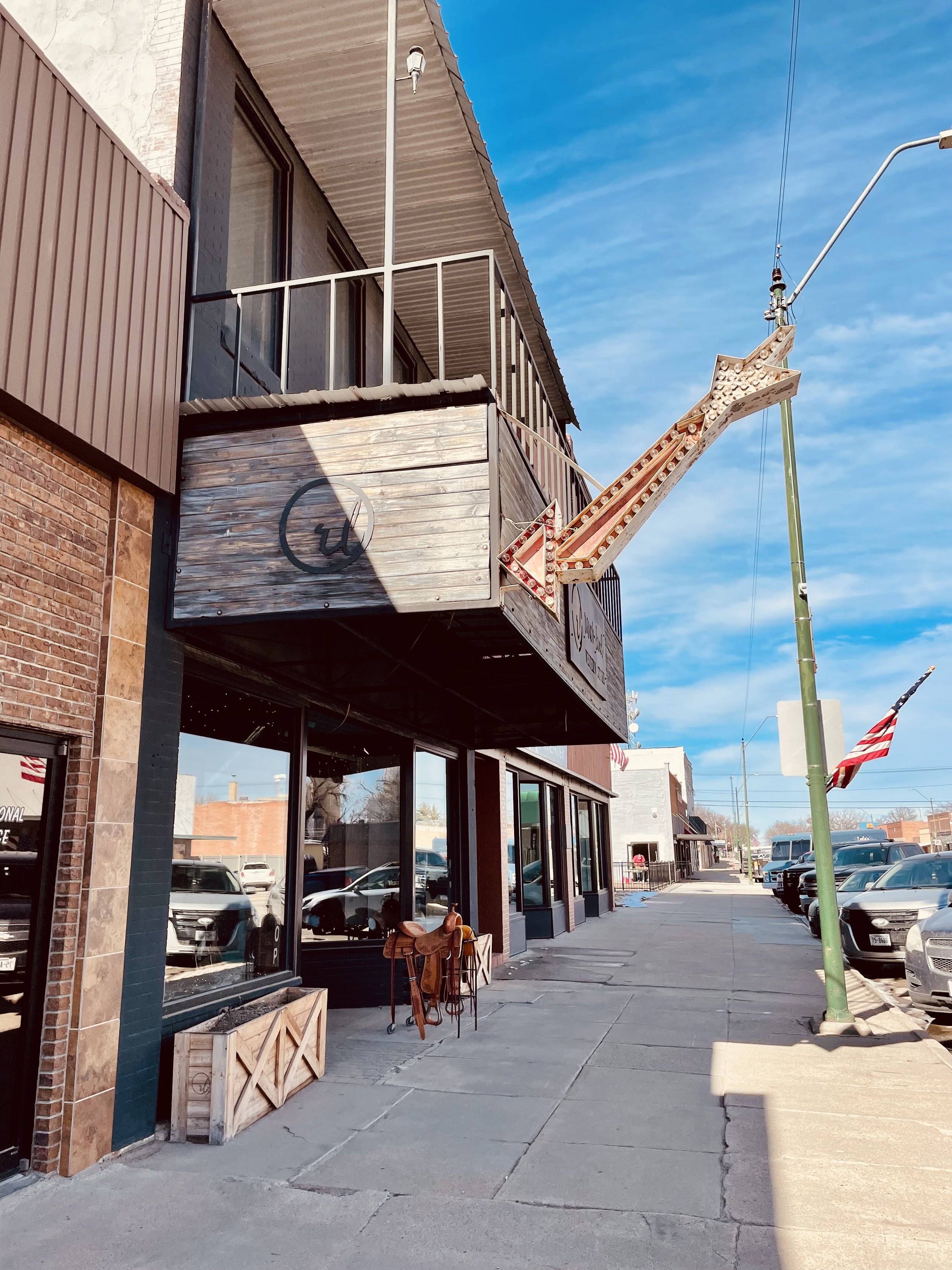 Empty sidewalk in front of a storefront with large windows, a hanging sign with a logo, and decorative wooden planters. There are parked cars and American flags in the distance under a blue sky.