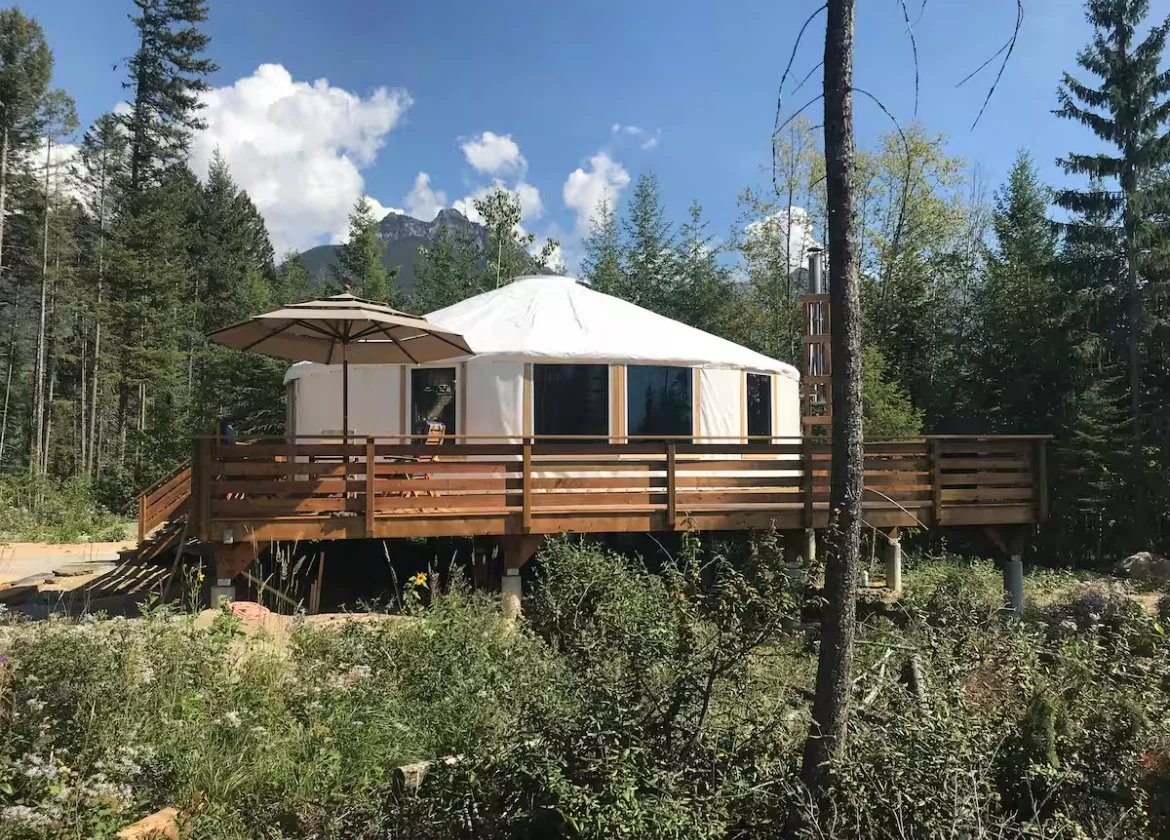A yurt with a wooden deck and umbrella, set in a forested area with mountains in the background.