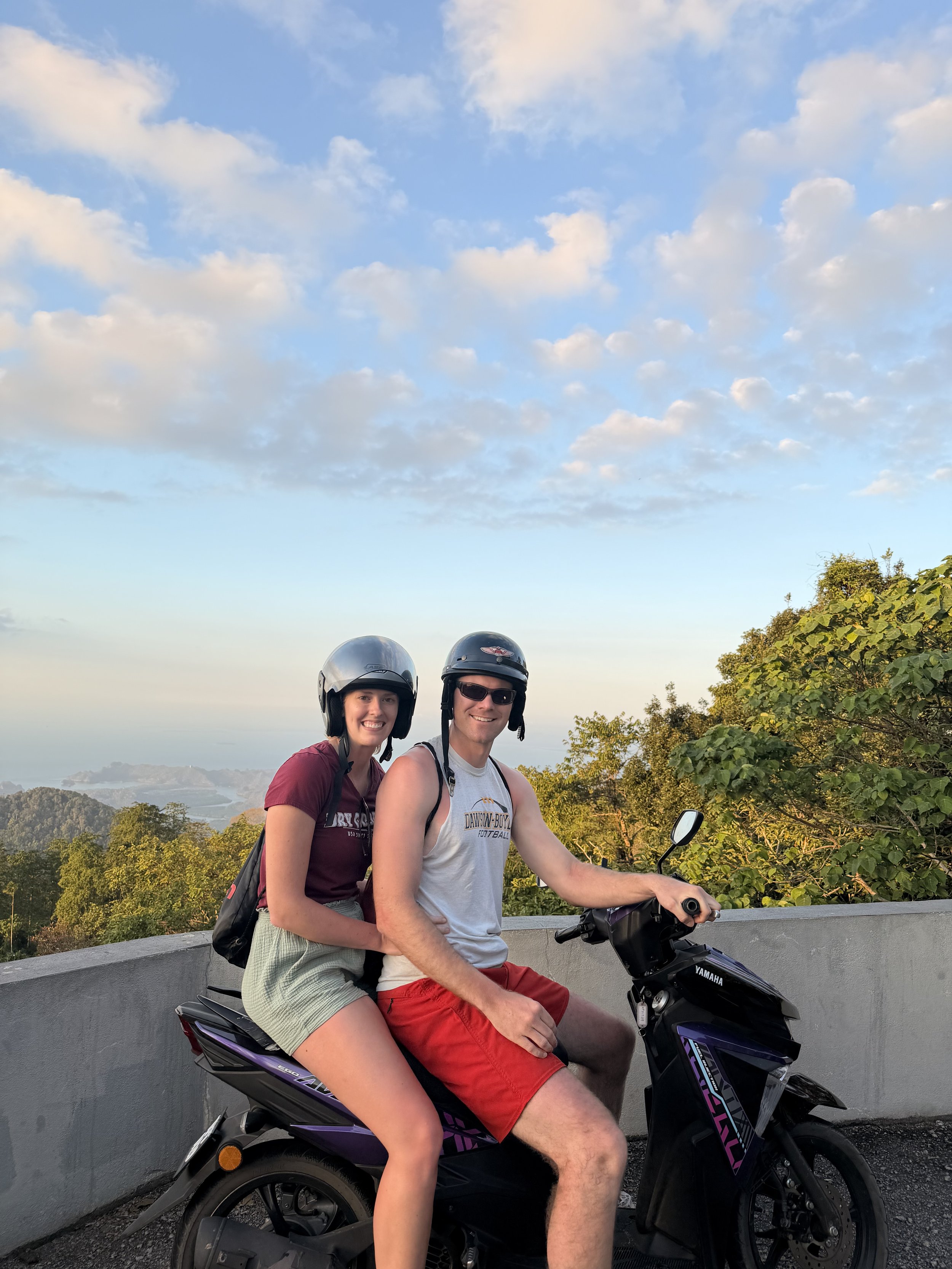 A young woman and man sitting on a purple and black Yamaha scooter, both wearing helmets and smiling at the camera outdoors. The woman is wearing a maroon shirt and shorts, and the man is wearing a white tank top and red shorts. There are trees and a scenic landscape in the background under a partly cloudy sky.