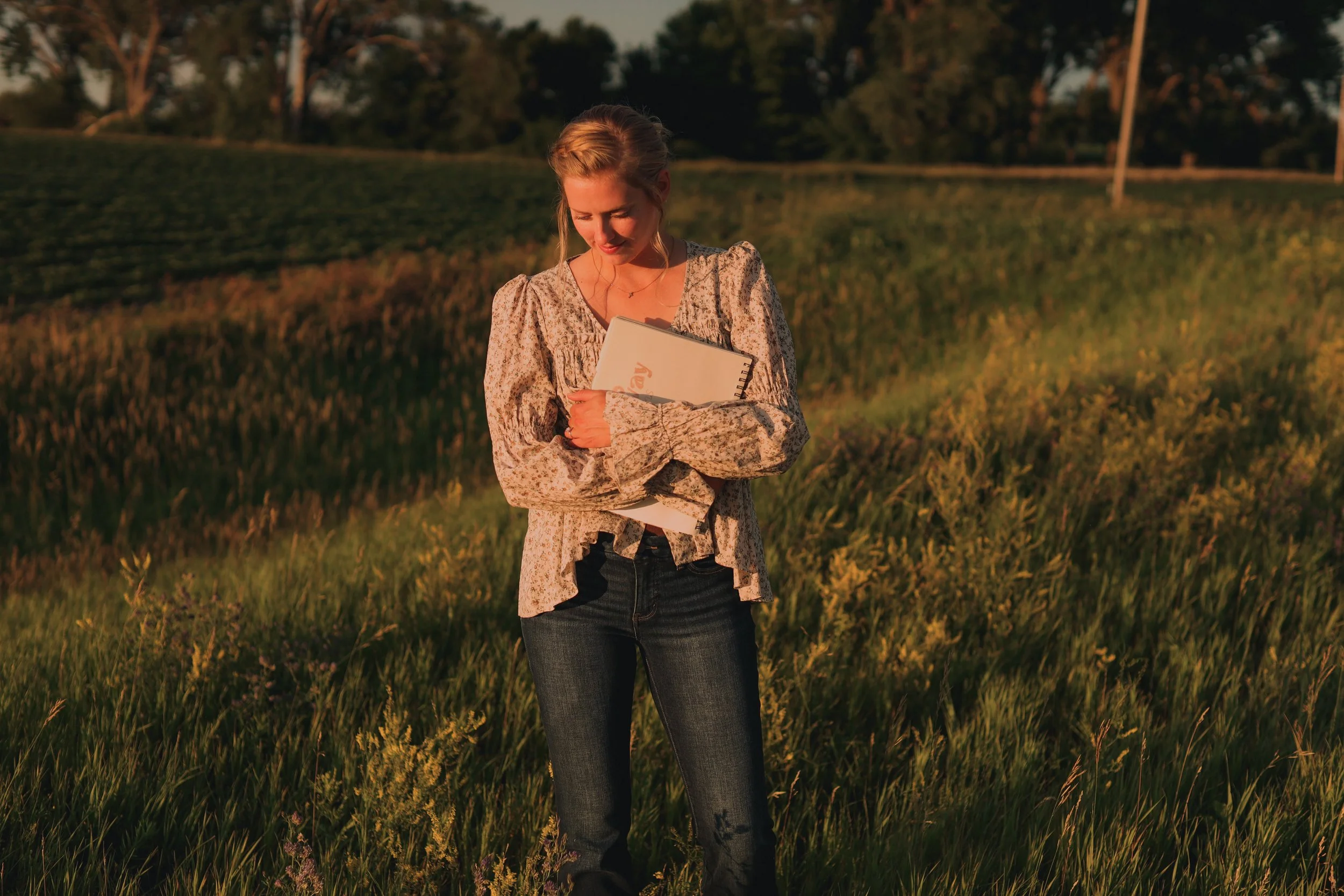 A woman with blonde hair wearing a floral blouse and jeans, holding a notebook, standing in a grassy field at sunset.
