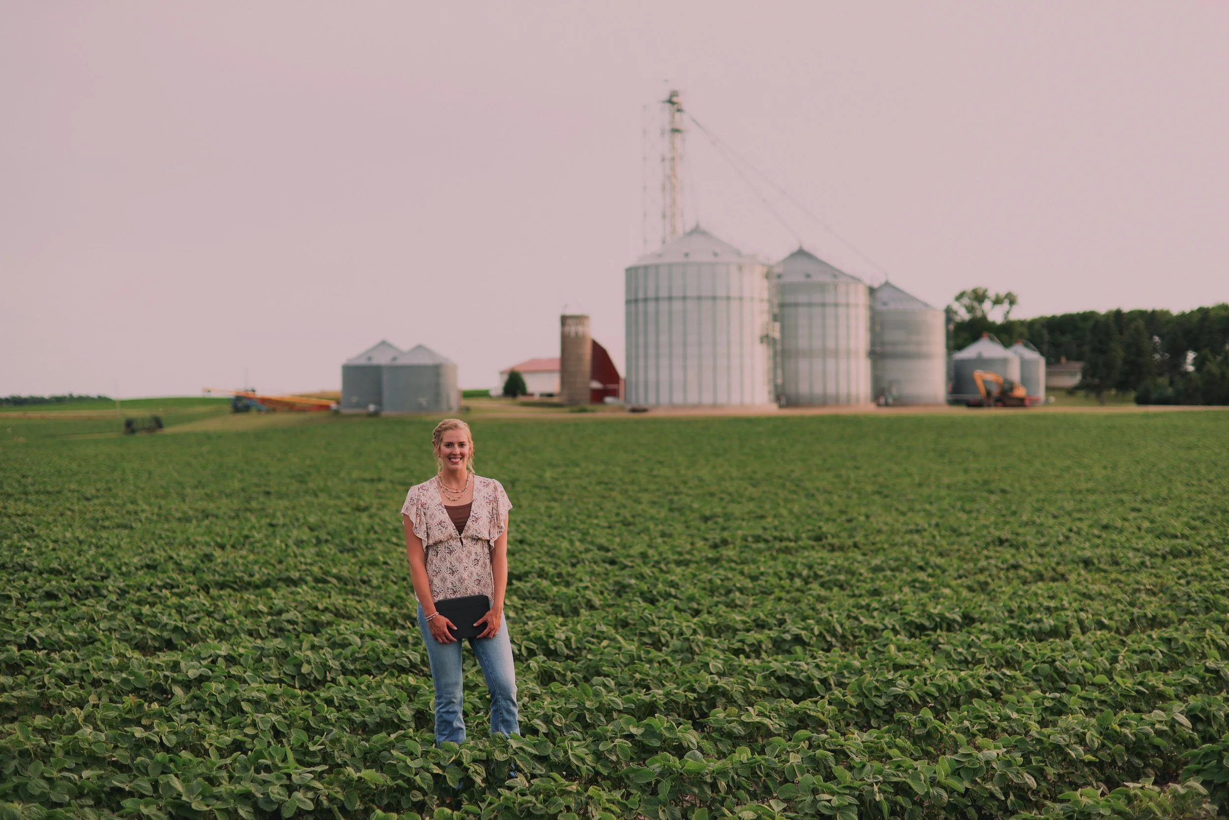 A woman standing in a field of green plants with farm silos and a red barn in the background, under a pink sky.