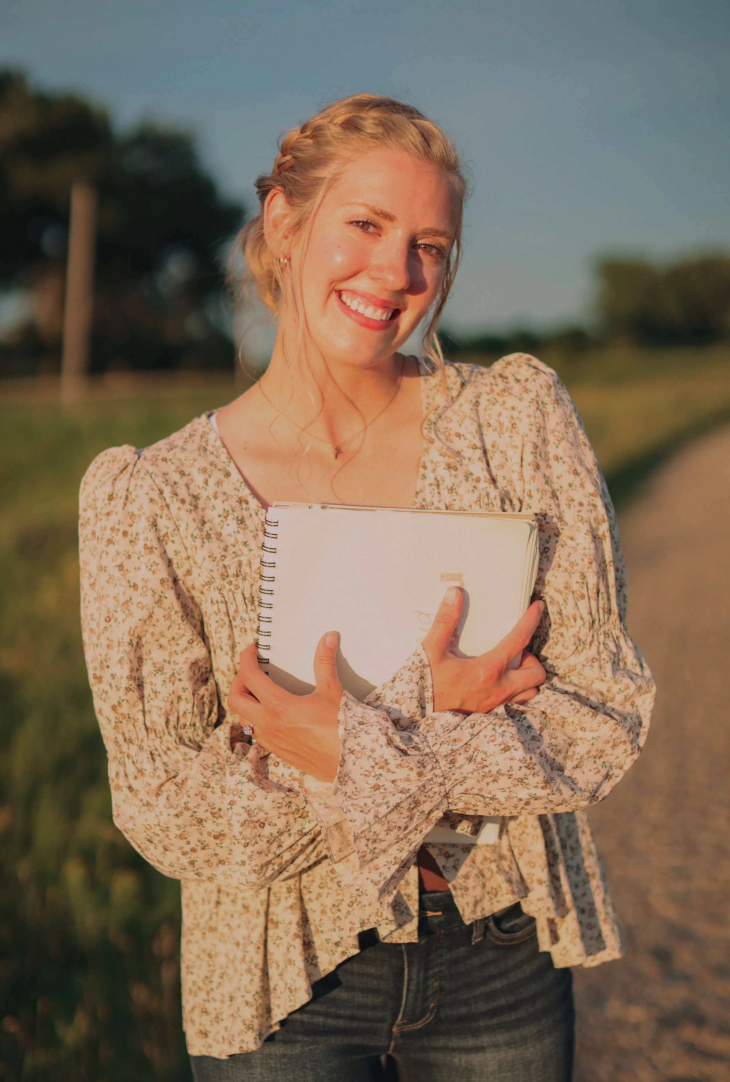 A smiling woman with blonde hair braided at the crown, wearing a floral blouse, holding a notebook, standing outdoors during sunset with trees in the background.