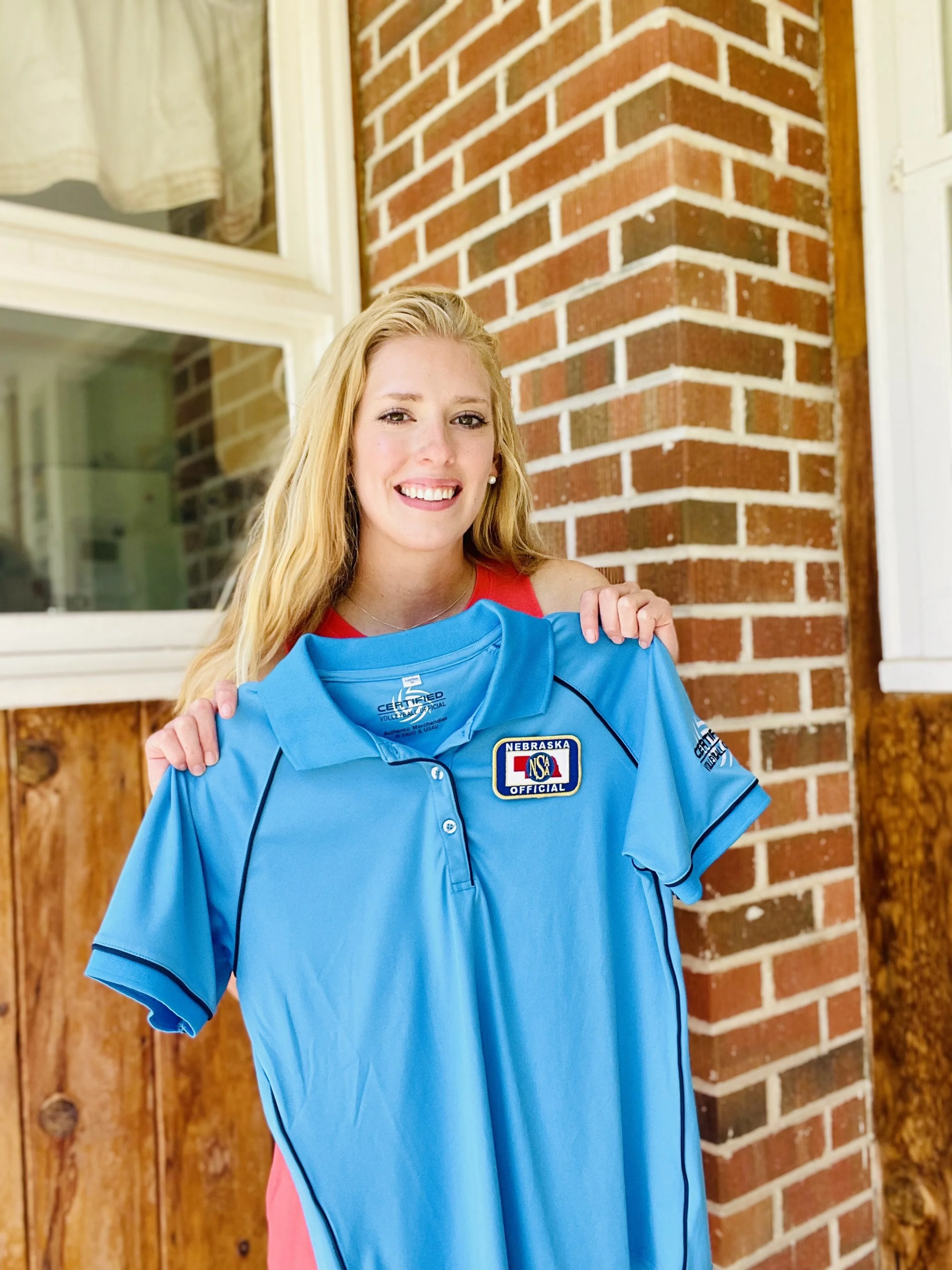 A young woman with blonde hair, wearing a red top and a blue sports jersey with an 'Official' badge, standing outside near a red brick wall.