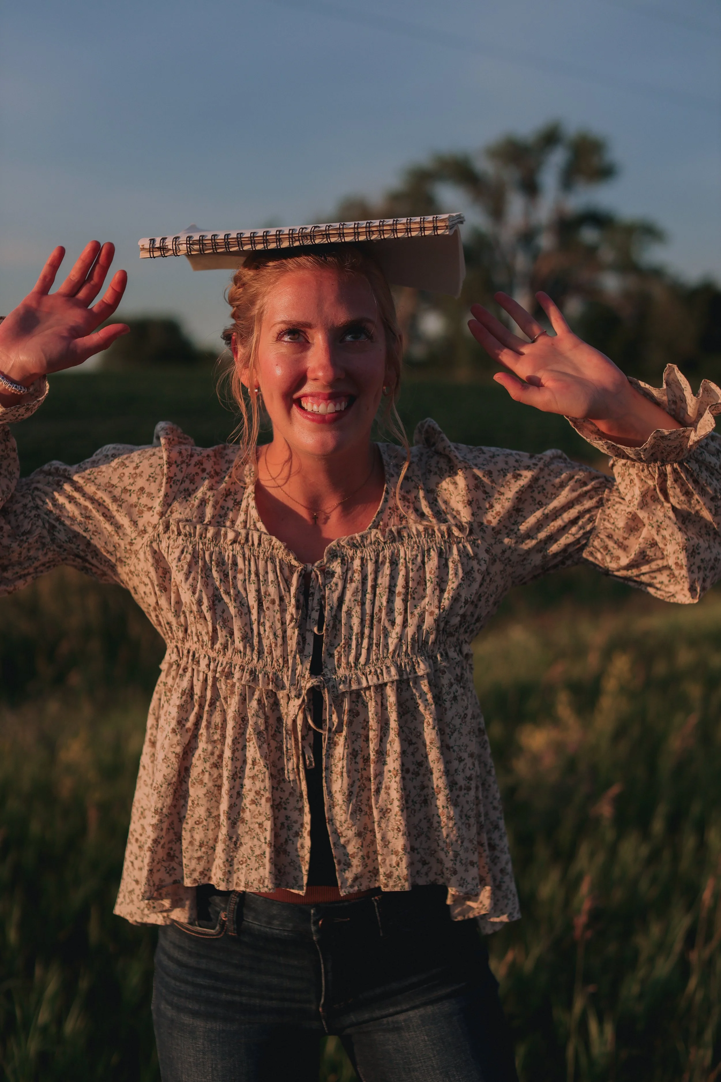 Young woman with blonde hair holding a notebook on her head outdoors in a grassy field at sunset, smiling and raising her hands.