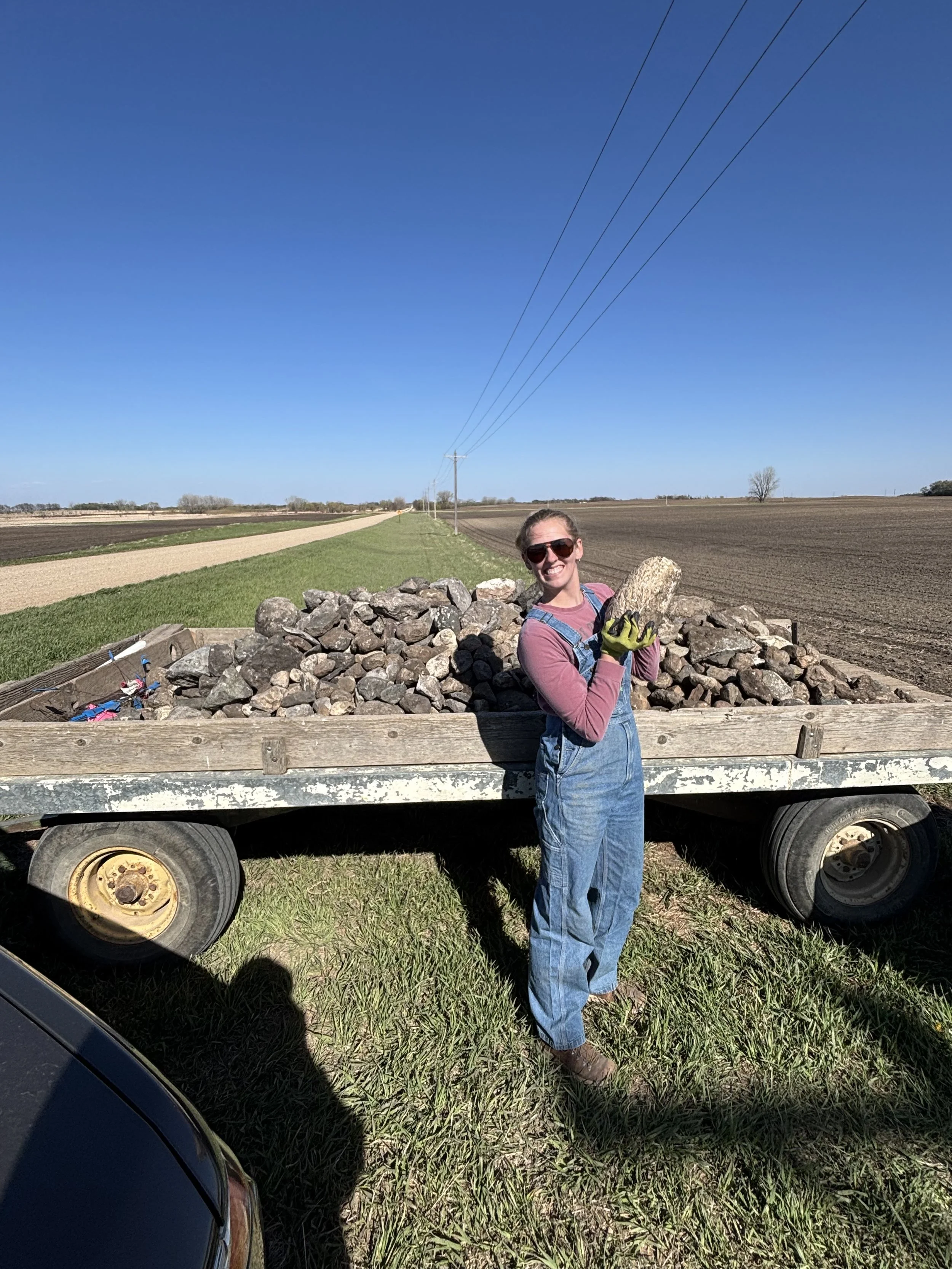 A woman wearing sunglasses, a pink shirt, and denim overalls, standing next to a flatbed truck filled with rocks, in a rural area with clear blue skies and open farmland in the background.