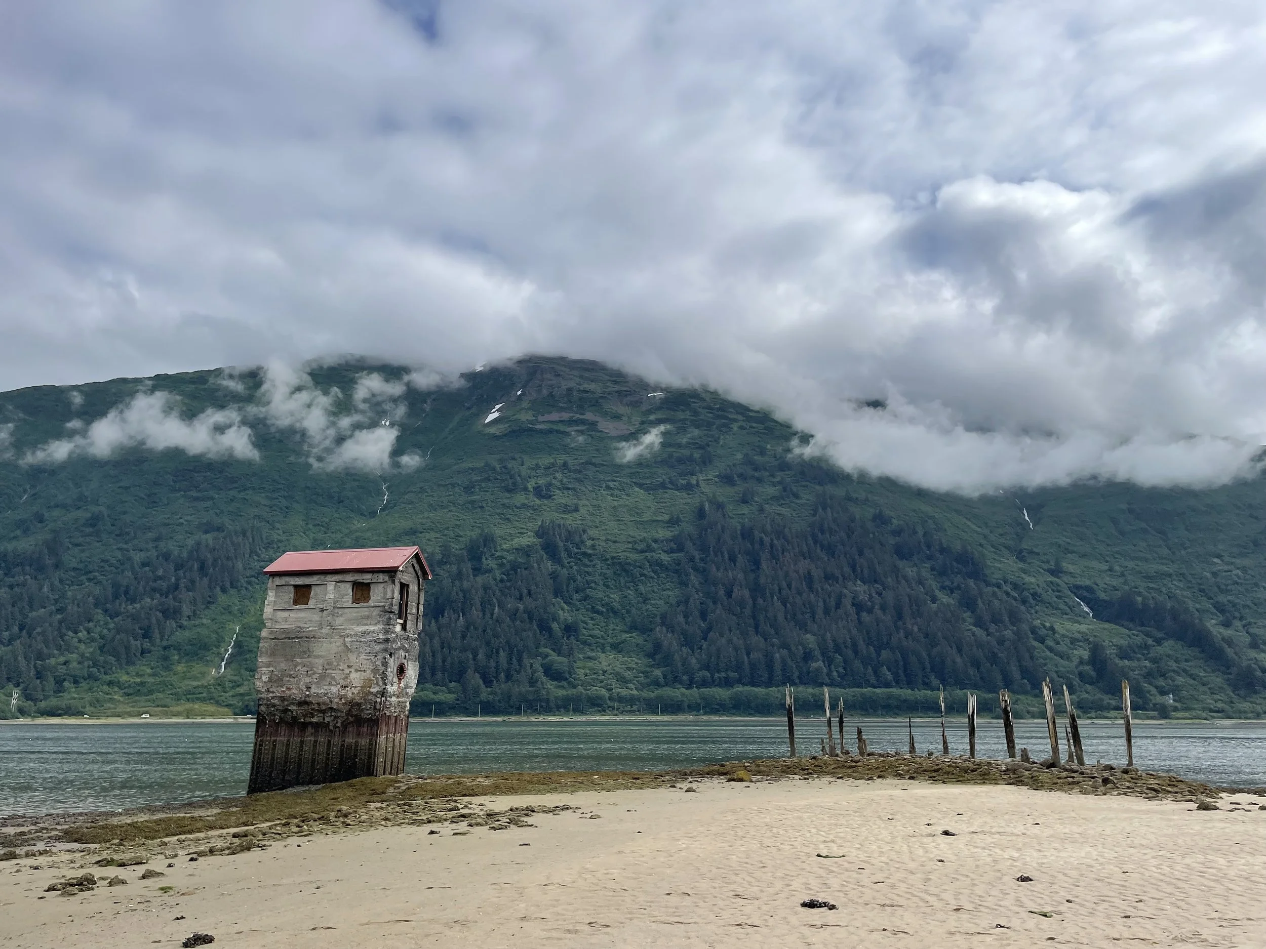 A stone tower with a red roof by a body of water, with lush green mountains and clouds in the background.