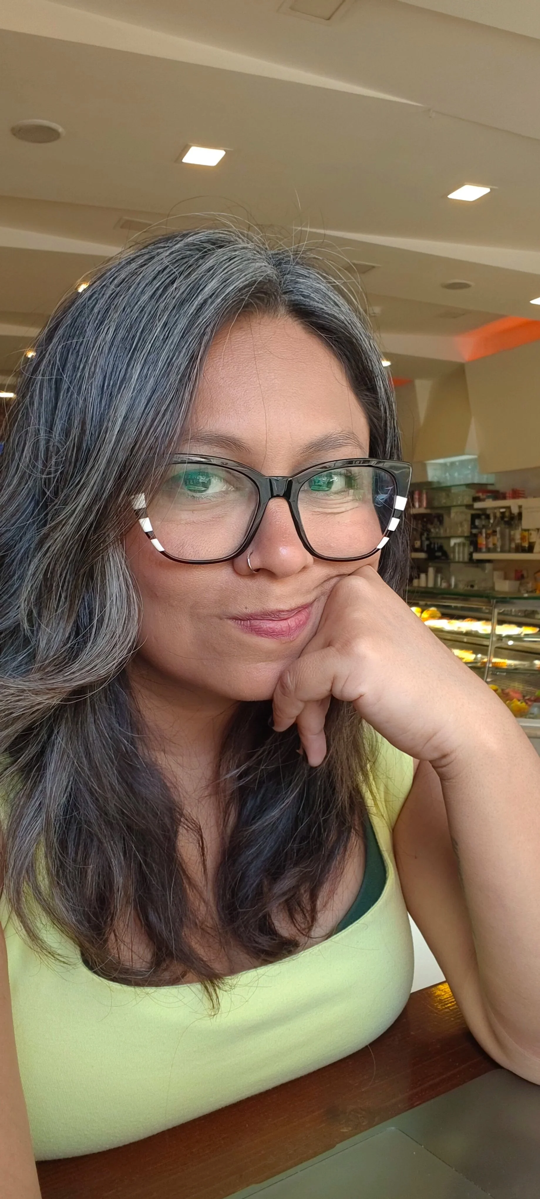 Una mujer con cabello gris y oscuro, usando gafas grandes y un piercing en la nariz, posa en un ambiente interior con una sonrisa sutil, con una tienda o mostrador de alimentos de fondo.