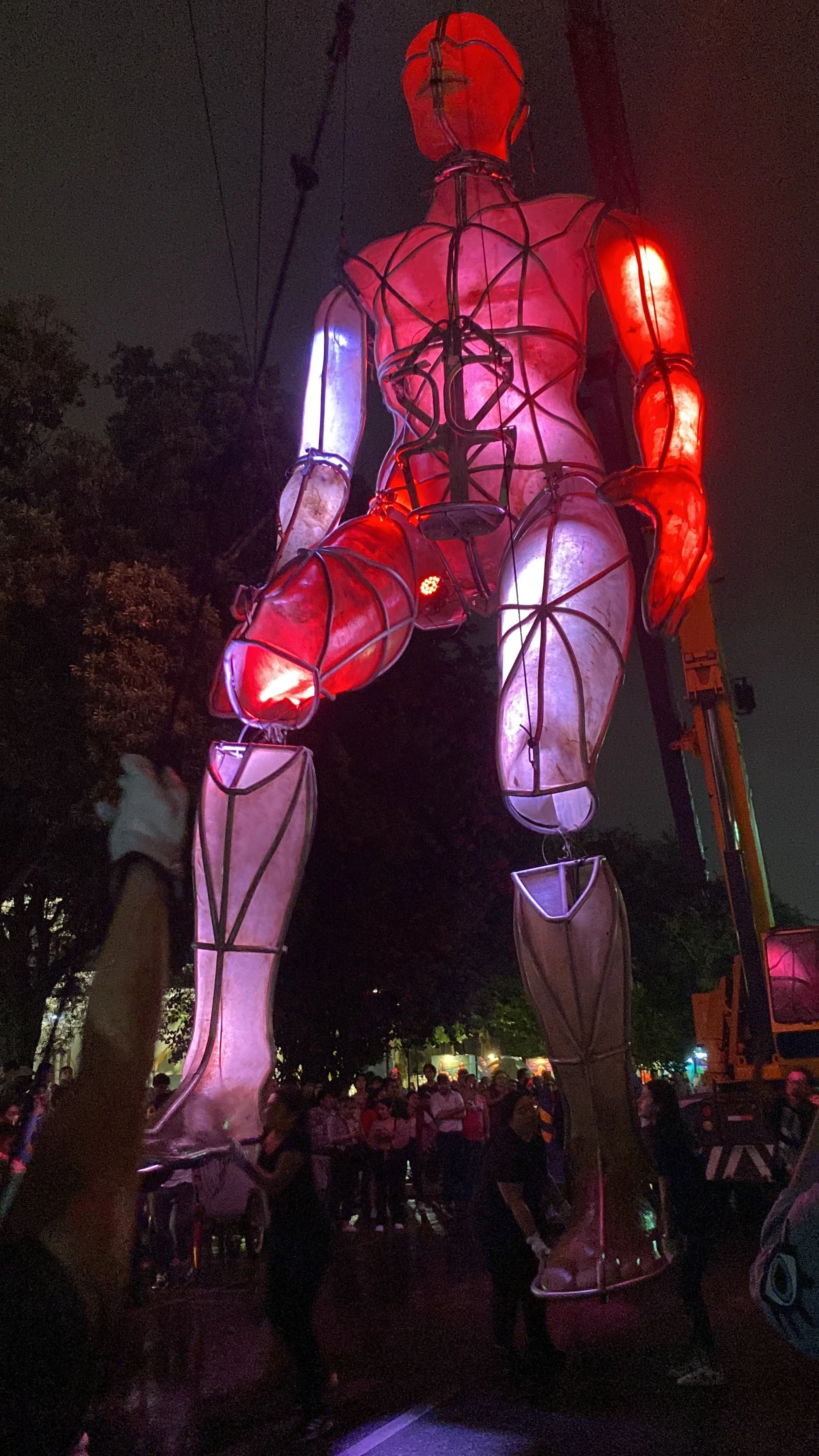 Escultura gigante iluminada de color rojo y blanco en forma de figura humana, ubicada en una feria nocturna con muchas personas alrededor y árboles en el fondo.