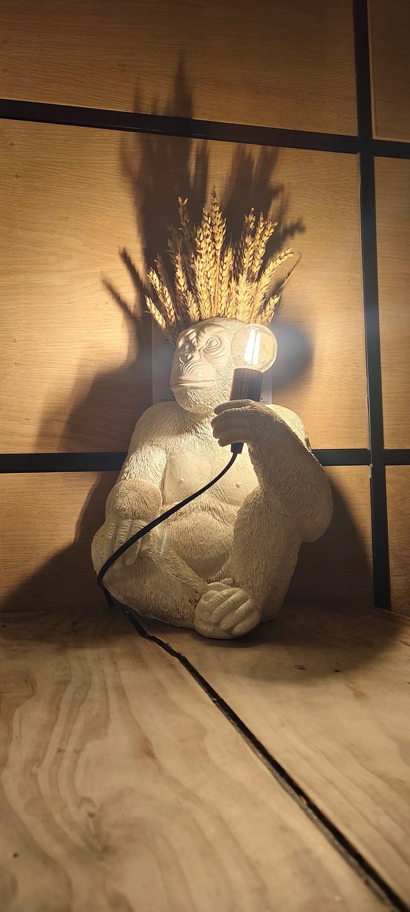 Estatua decorativa de un gorila sosteniendo una bombilla, con una cabeza decorada con espigas de trigo y sombra en la pared de fondo.