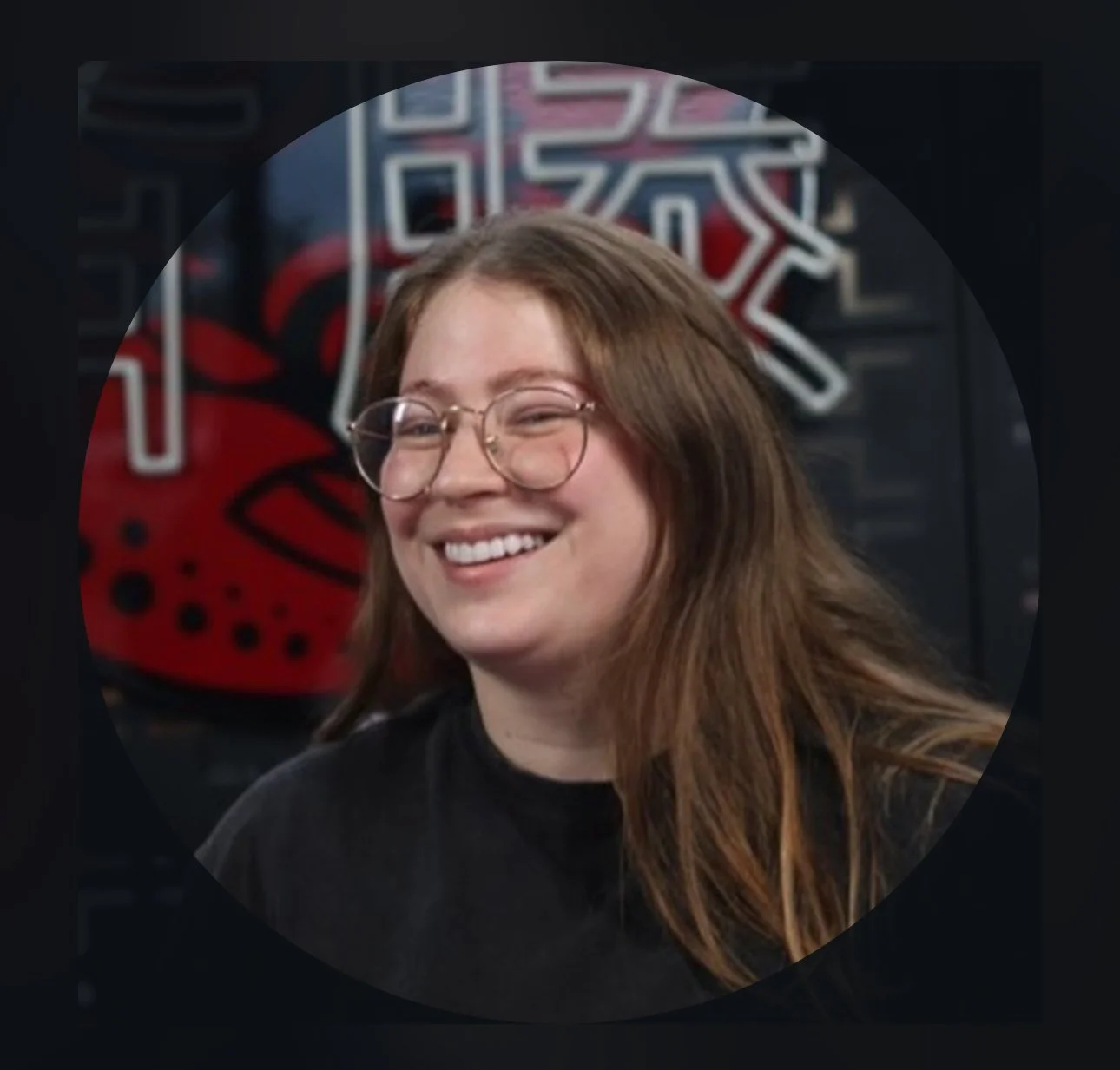 A female tattoo artist with long reddish-brown hair, glasses, and a black shirt, smiling in front of a neon red and white sign.