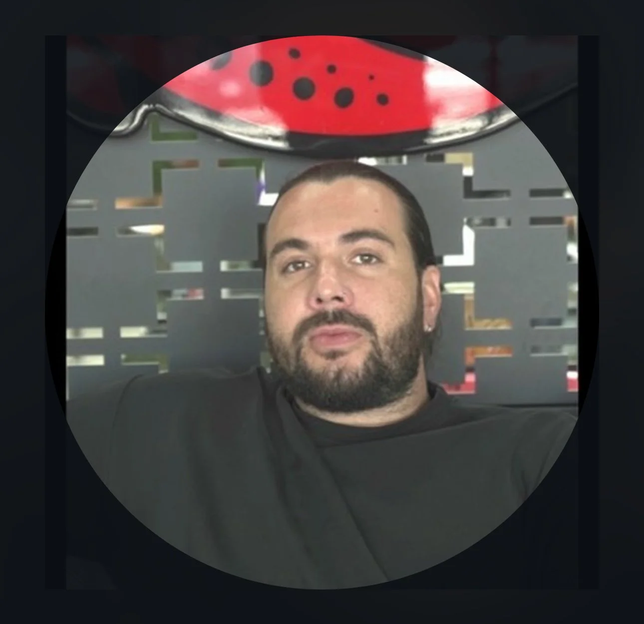A male tattoo artist with dark hair and a beard, sitting on a black sofa, with a gray decorative partition behind him and a decorative object with red and black colors above his head.
