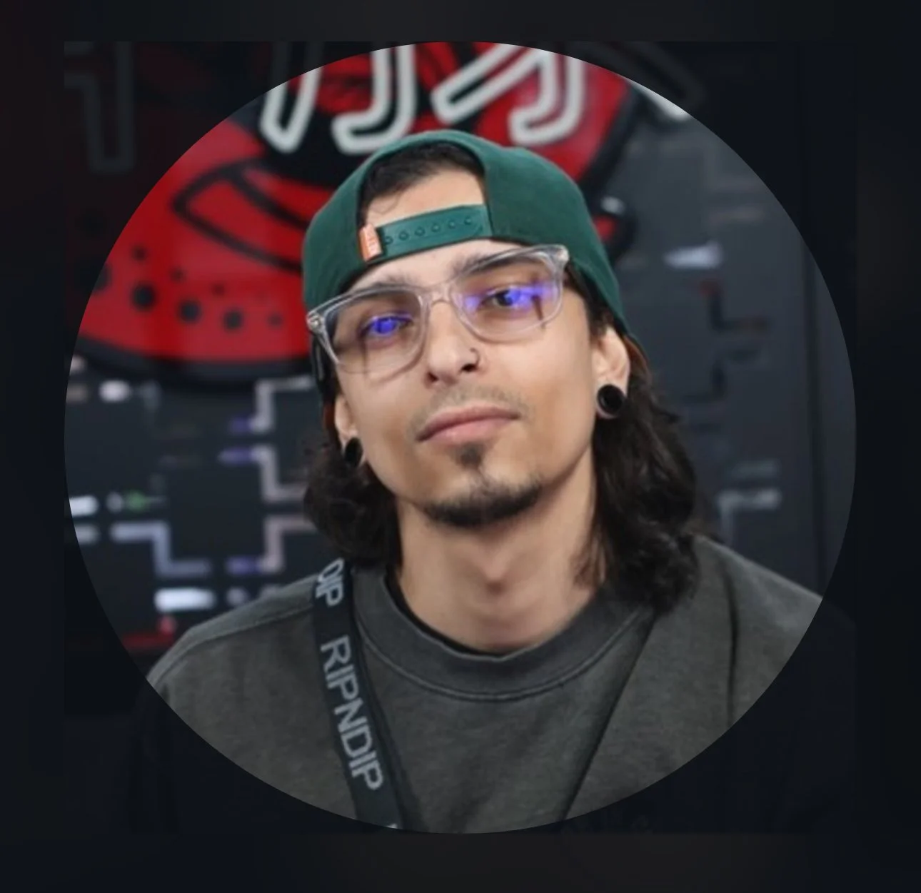 Tattoo Artist with long dark hair, glasses, a nose ring, and black earrings, wearing a green baseball cap backwards and a dark gray shirt. He has a lanyard with that says 'RIPNDIP' around his neck, standing in front of a black and red background.