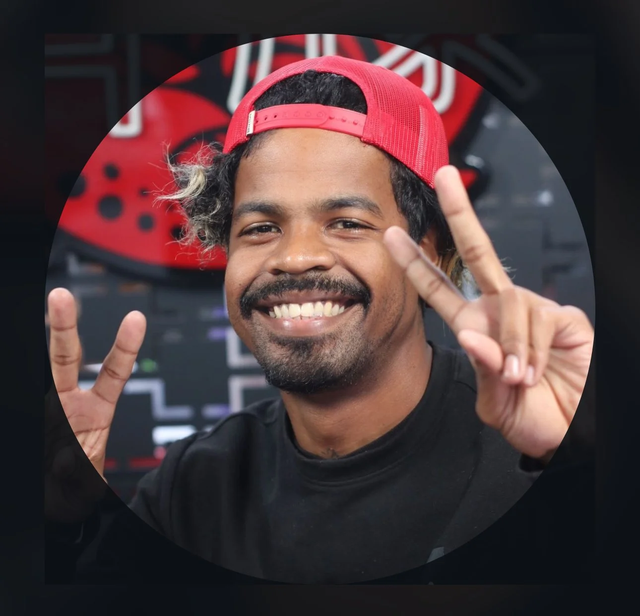 A smiling male tattoo artist with a beard and mustache, wearing a red cap backwards, making peace signs with both hands.