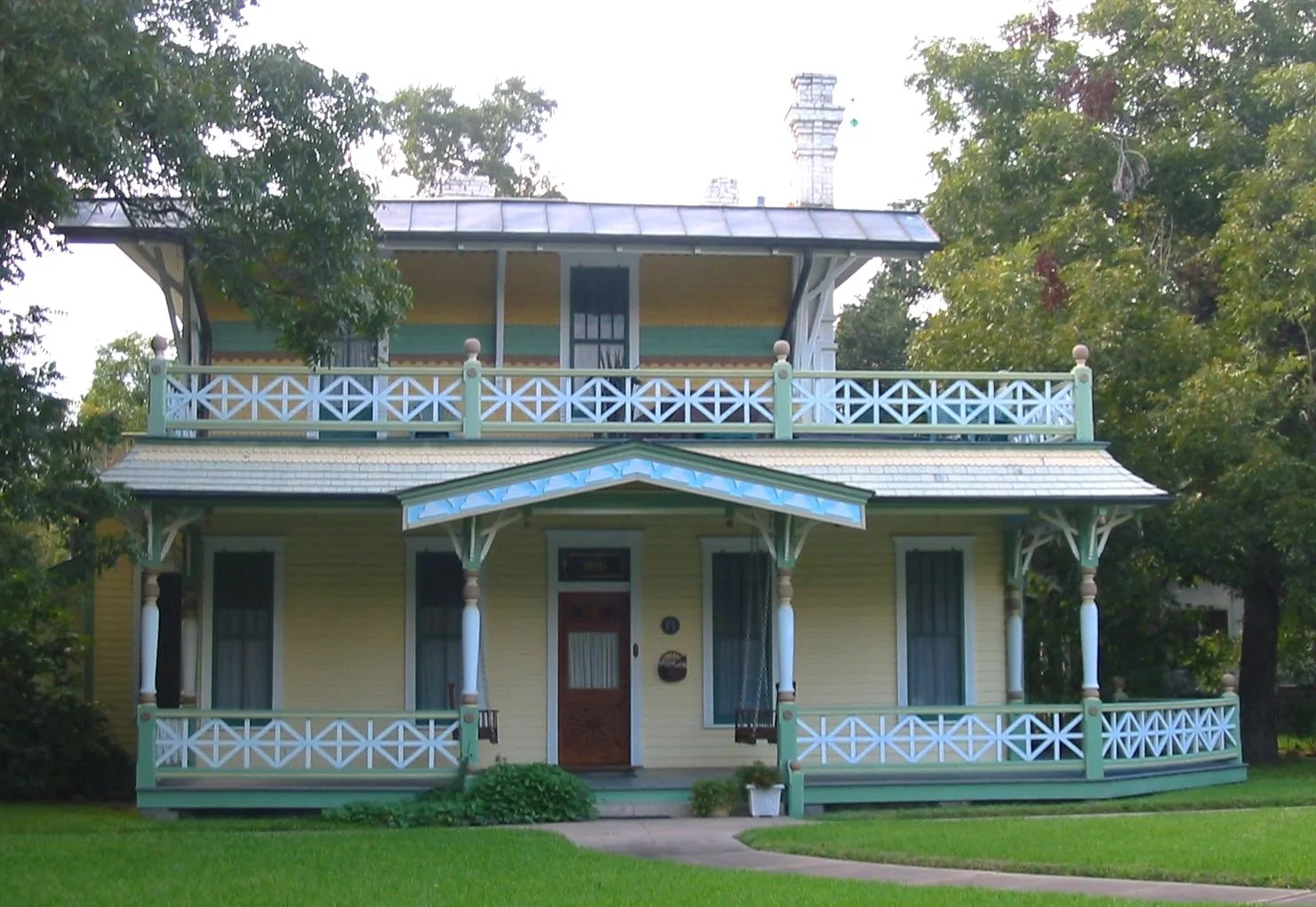Colorful two-story house with a wraparound porch, decorative railings, and a small balcony on the second floor, surrounded by greenery.