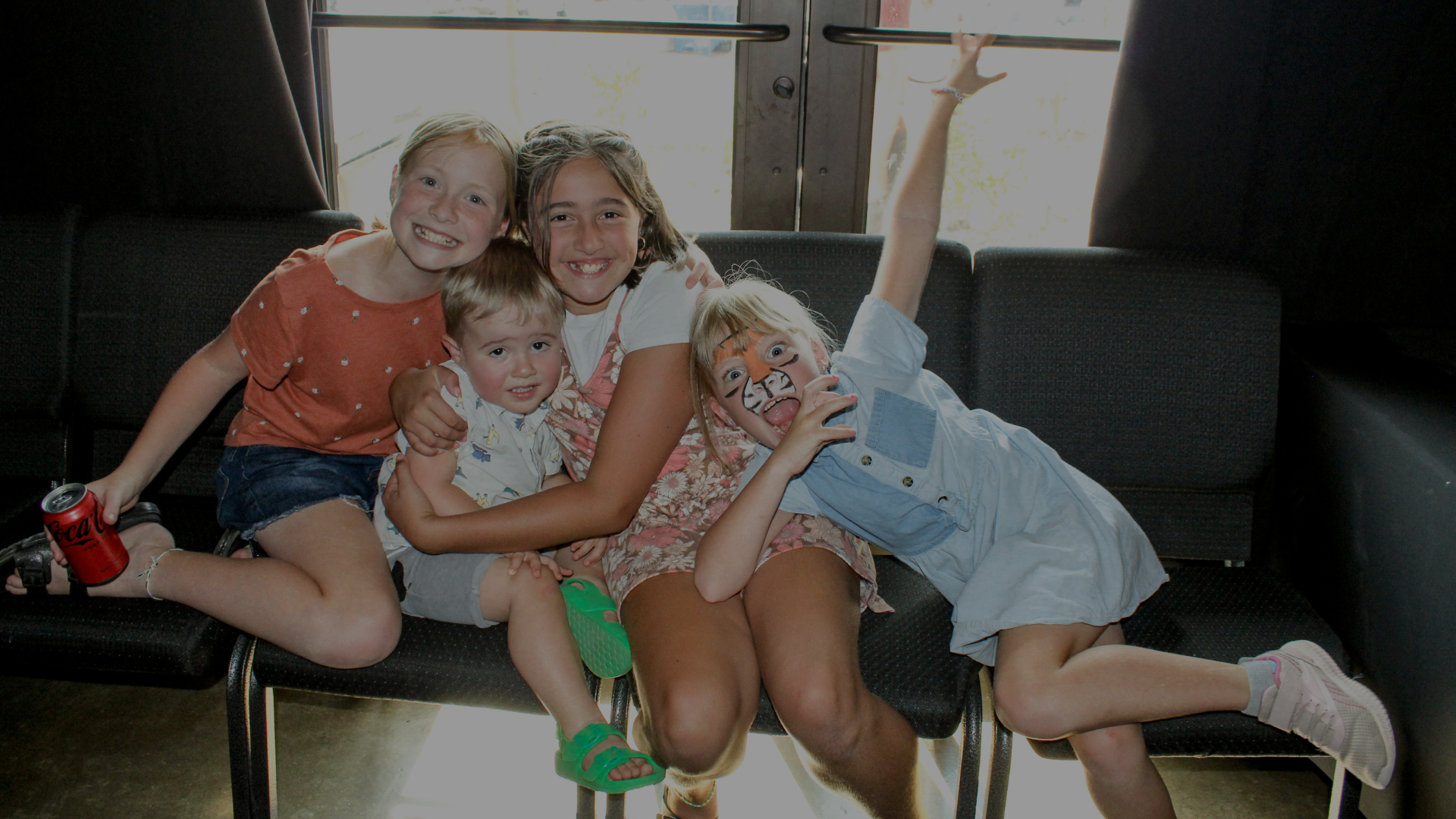Group of five children sitting on a black couch, smiling and posing for the camera. One girl has tiger face paint and is lying on her side, holding her face with her hand, with kids hugging her. Another girl has her arm raised above her head, holding onto a metal bar. One boy is holding a soda can. They are in a room with large windows in the background.