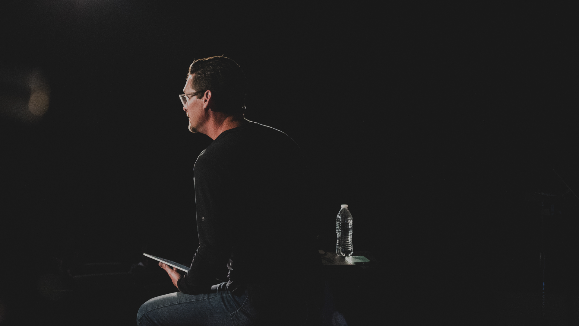 A man with glasses sitting in a dark room, holding a tablet, with a water bottle on a small table beside him.