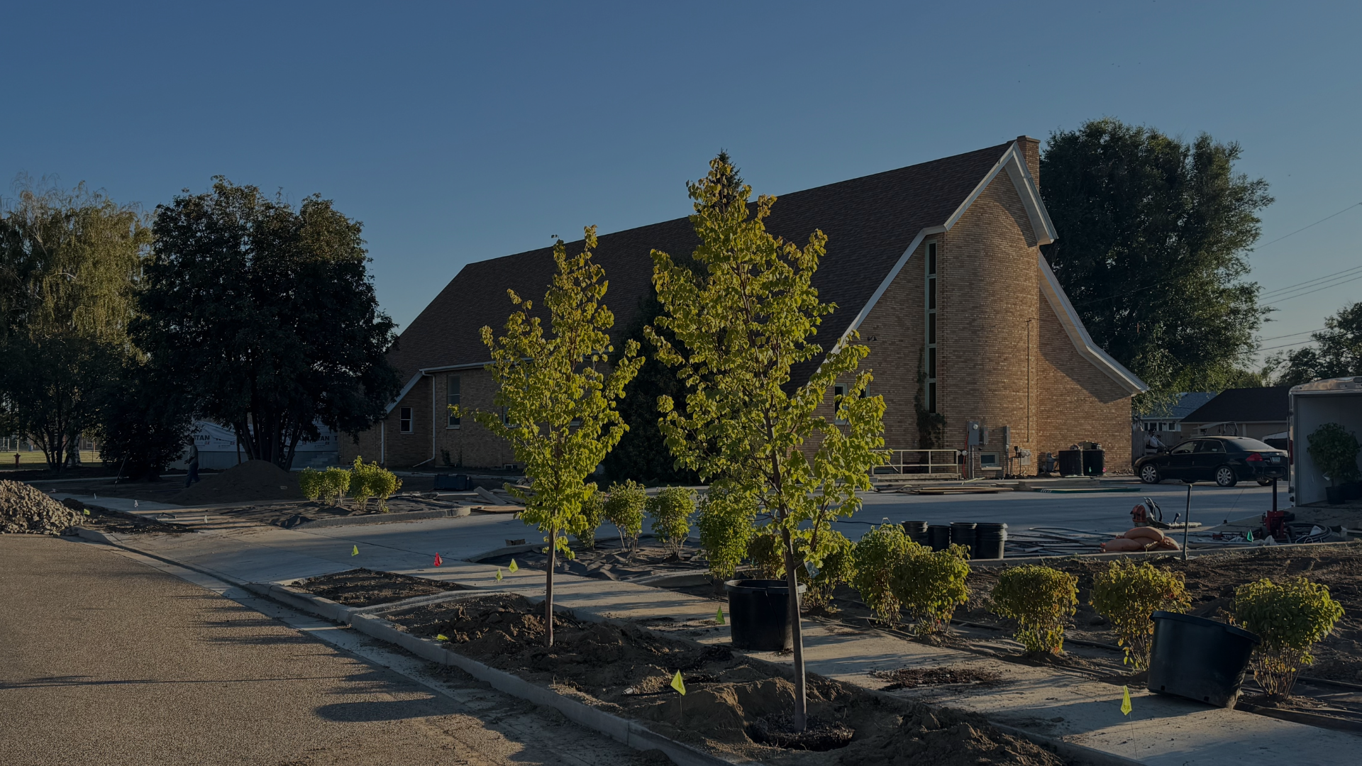 A church under construction with trees and plants in the foreground, construction materials and vehicles nearby, and a clear blue sky.