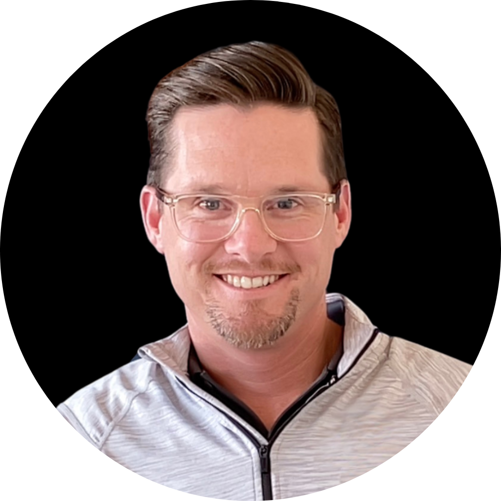 A man with glasses and a short beard, smiling, wearing a light gray zip-up sports jacket, against a black background.