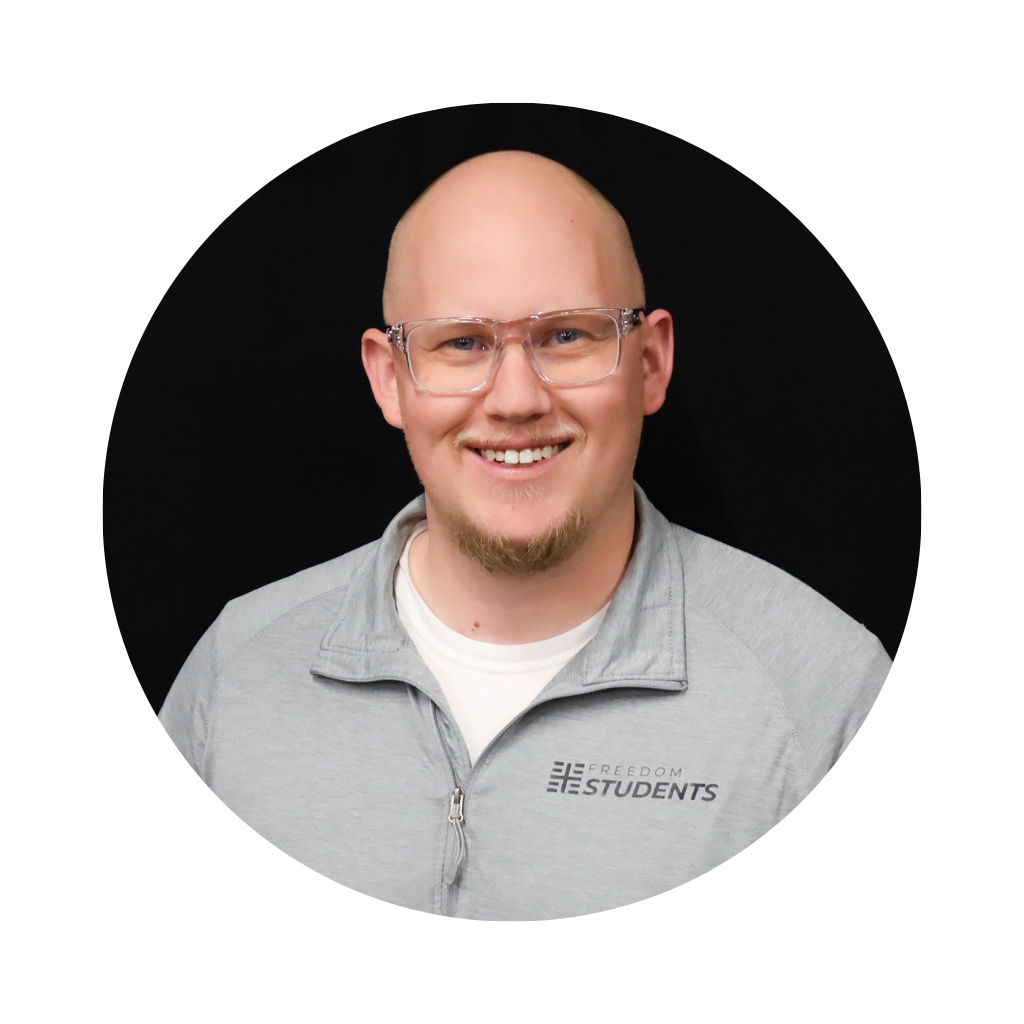 A smiling man with glasses and a light beard wearing a light gray zip-up jacket with 'FREEDOM STUDENTS' logo, standing against a black background.
